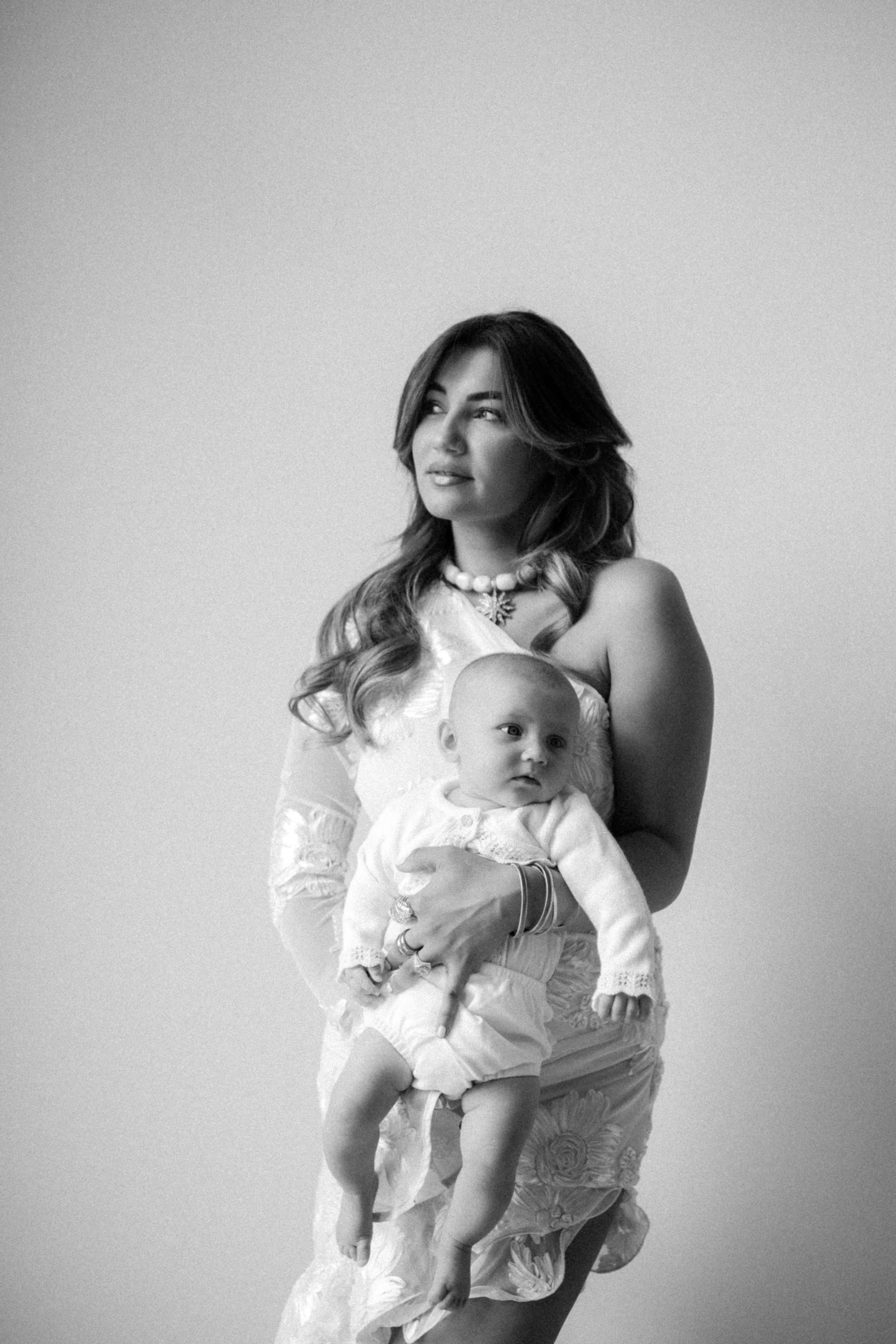 A woman holding a baby in her arms. The woman has long, wavy hair and is dressed in a floral outfit with jewelry, including a necklace and bracelets. The baby is wearing a light-colored outfit and looks to the side. The photo is in black and white.