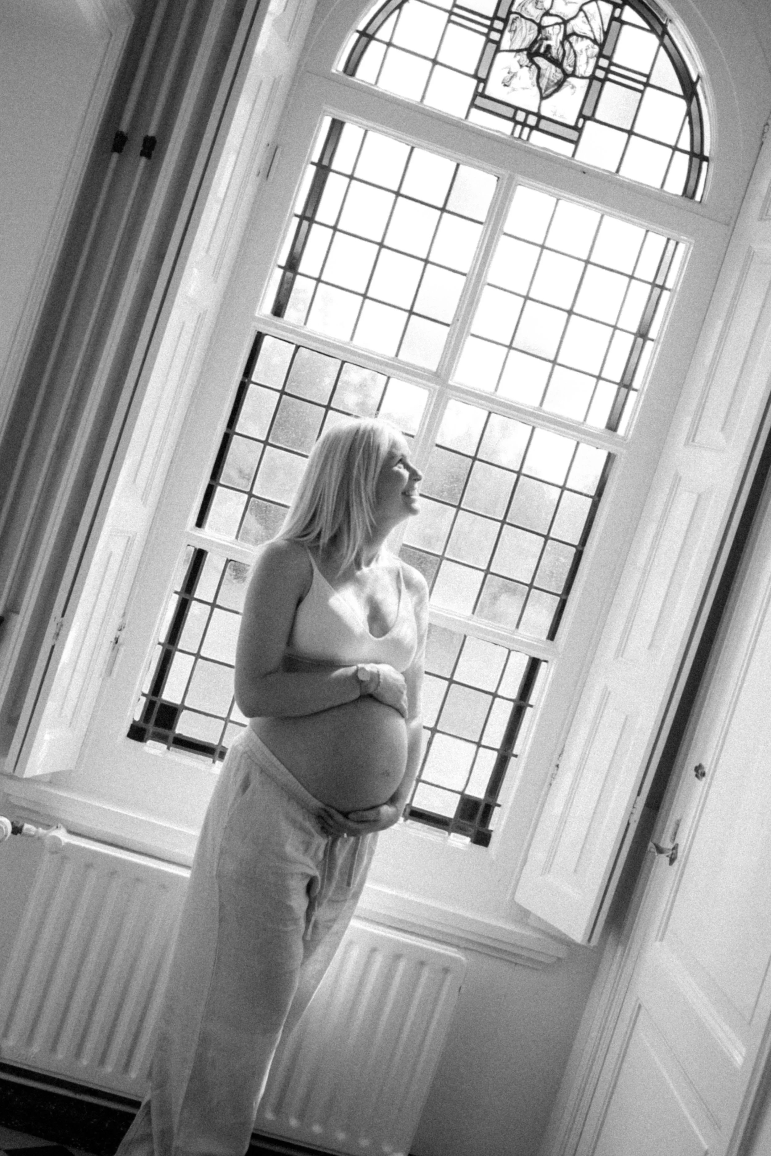 A pregnant woman in a tank top and sweatpants standing beside a large window with stained glass, smiling and looking out.