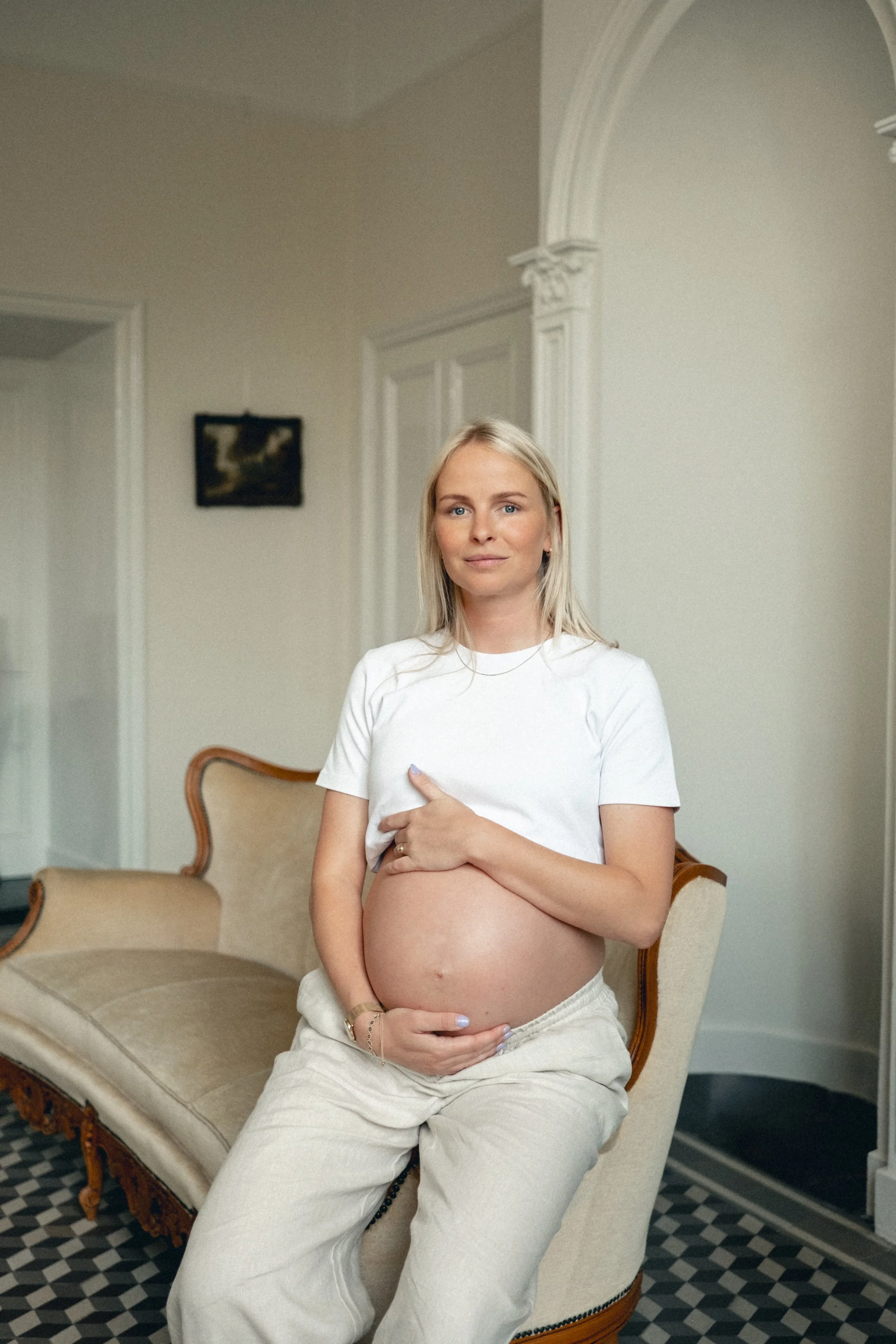 A pregnant woman sitting on a vintage beige sofa in a well-lit room, holding her baby bump with her hand, wearing a white t-shirt and cream-colored pants.