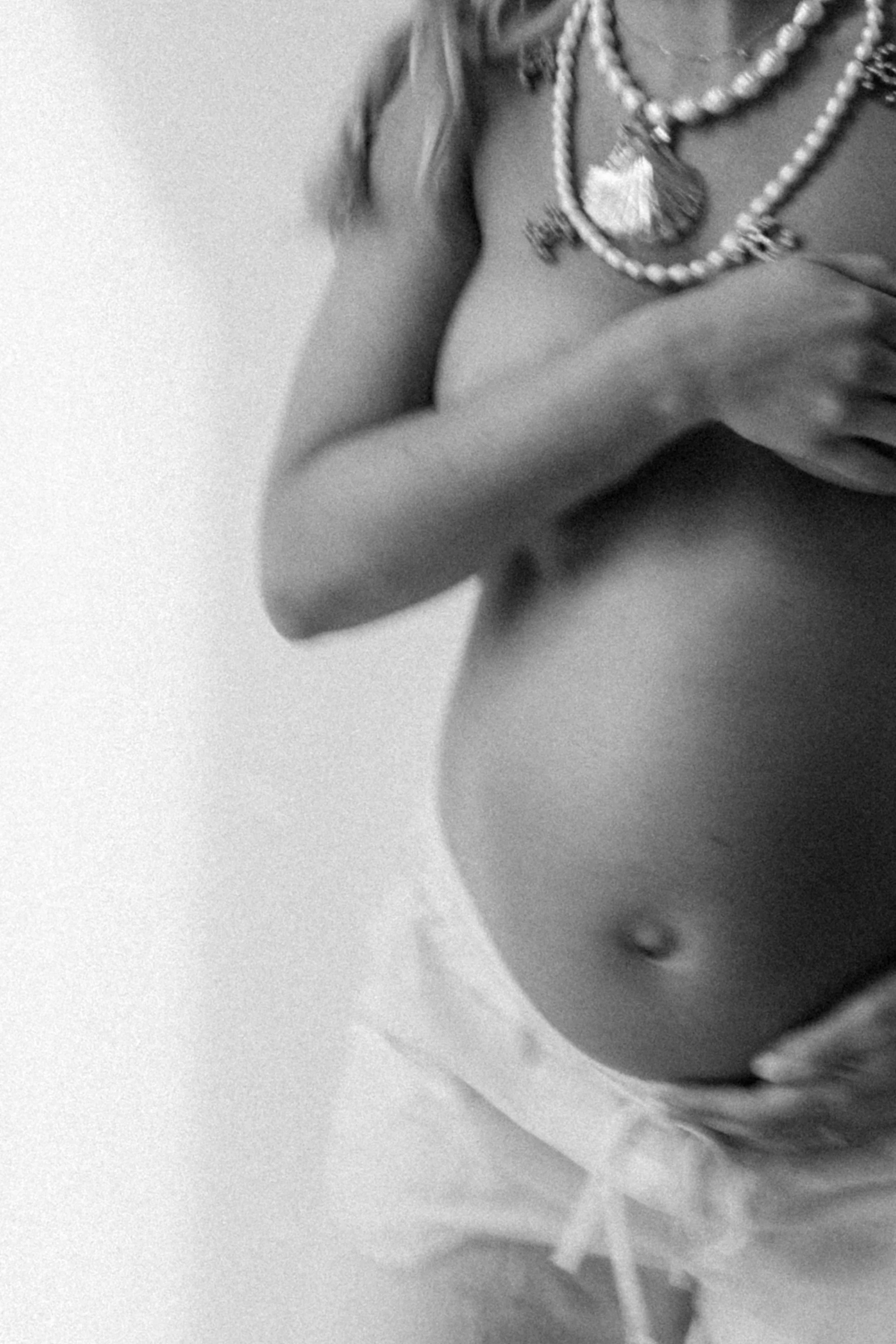 A pregnant woman wearing layered necklaces and cropped clothing, holding her belly with one hand and touching her shoulder with the other, in black and white.
