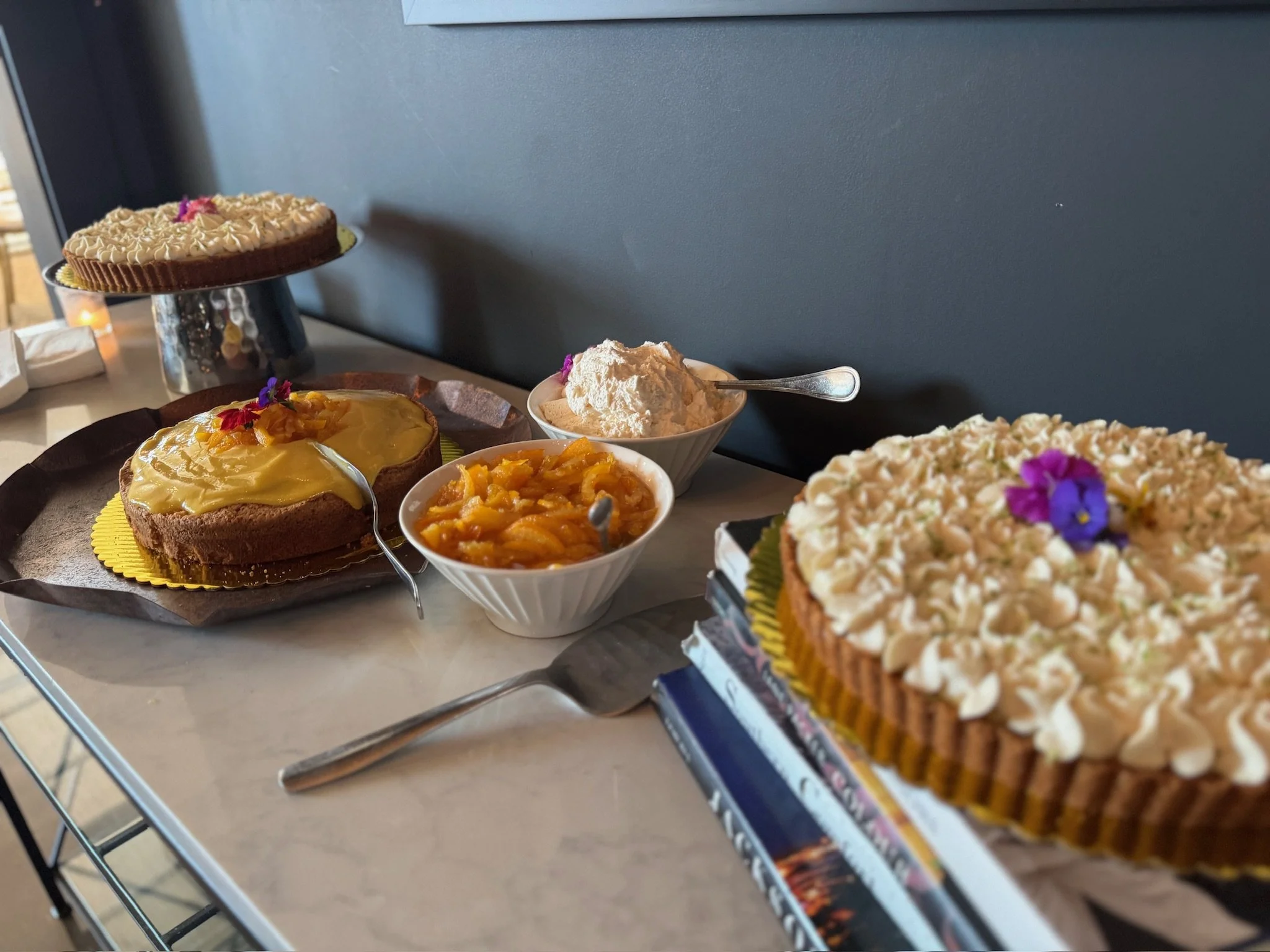 Assorted desserts including a peach tart with edible flowers, a lemon cake with fruit toppings, bowls of whipped cream and fruit compote, and a large decorated cake with purple flowers.