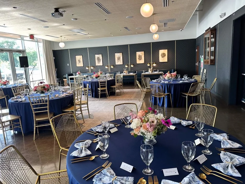 Elegant banquet room set for a formal event, with round tables draped in navy blue tablecloths, gold chairs, floral centerpieces, and place settings with glassware and silverware.