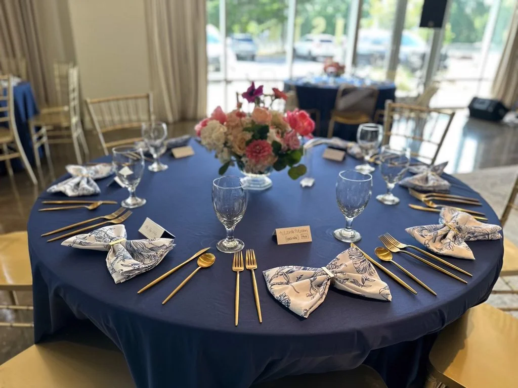 A round banquet table set with navy blue tablecloth, gold cutlery, water glasses, and floral napkins arranged as bows. A floral centerpiece with pink and white flowers is in the middle, with a banquet hall and large window in the background.