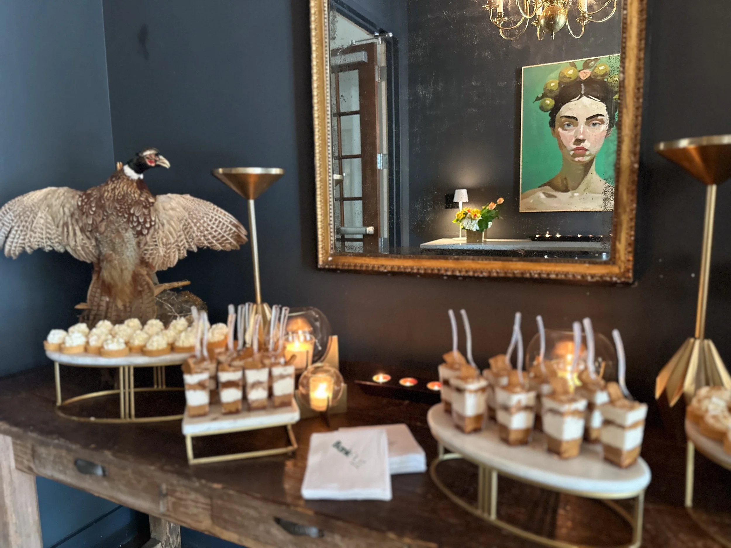 Display of assorted desserts on a wooden table with decorative items, including a model of a peacock, candles, and a framed portrait of a woman with a floral crown reflected in a mirror on a dark-colored wall.