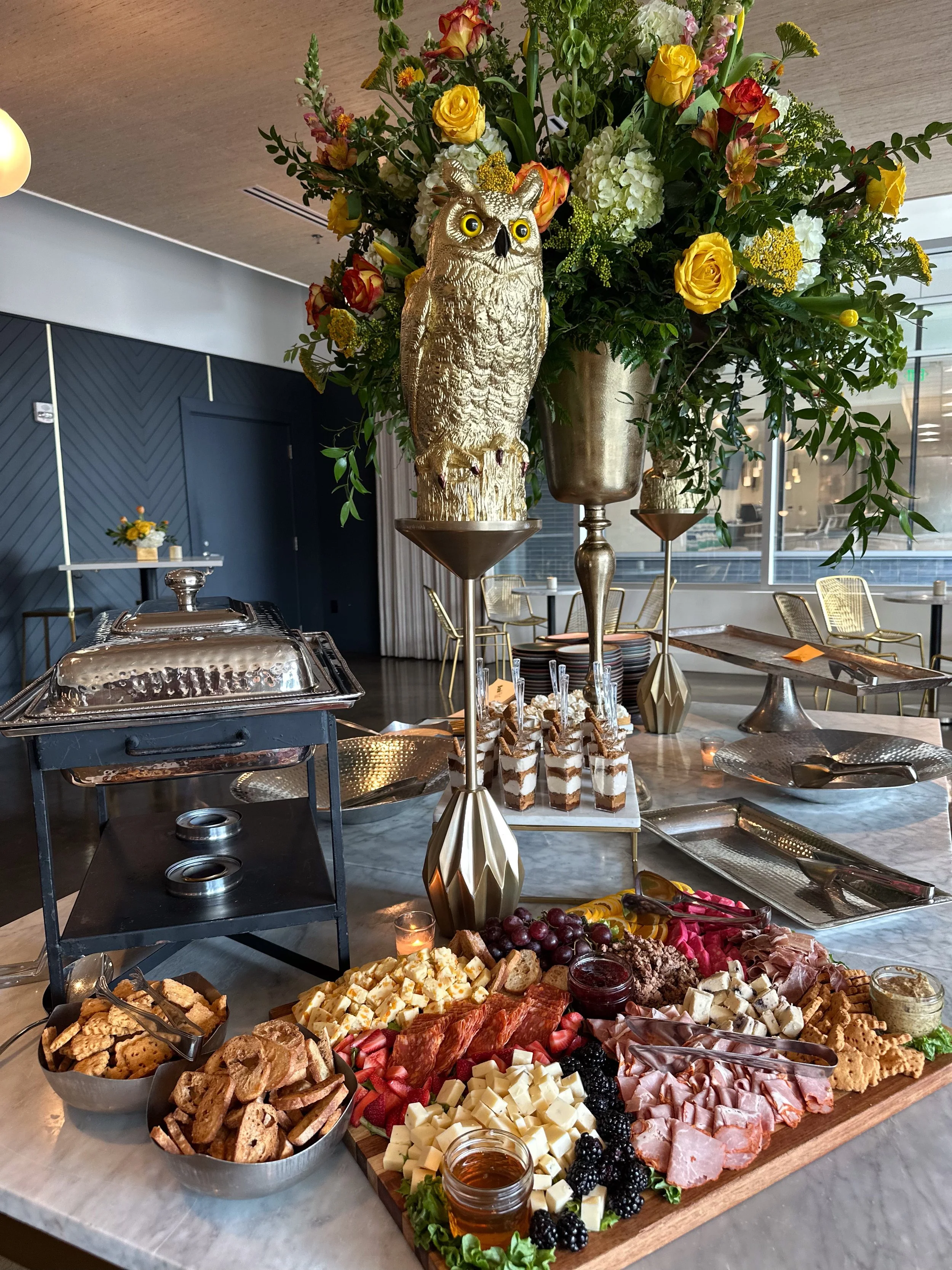 A cheese and charcuterie platter with grapes, berries, crackers, and honey in the foreground, topped with a large floral arrangement and a decorative gold owl sculpture on a buffet table.