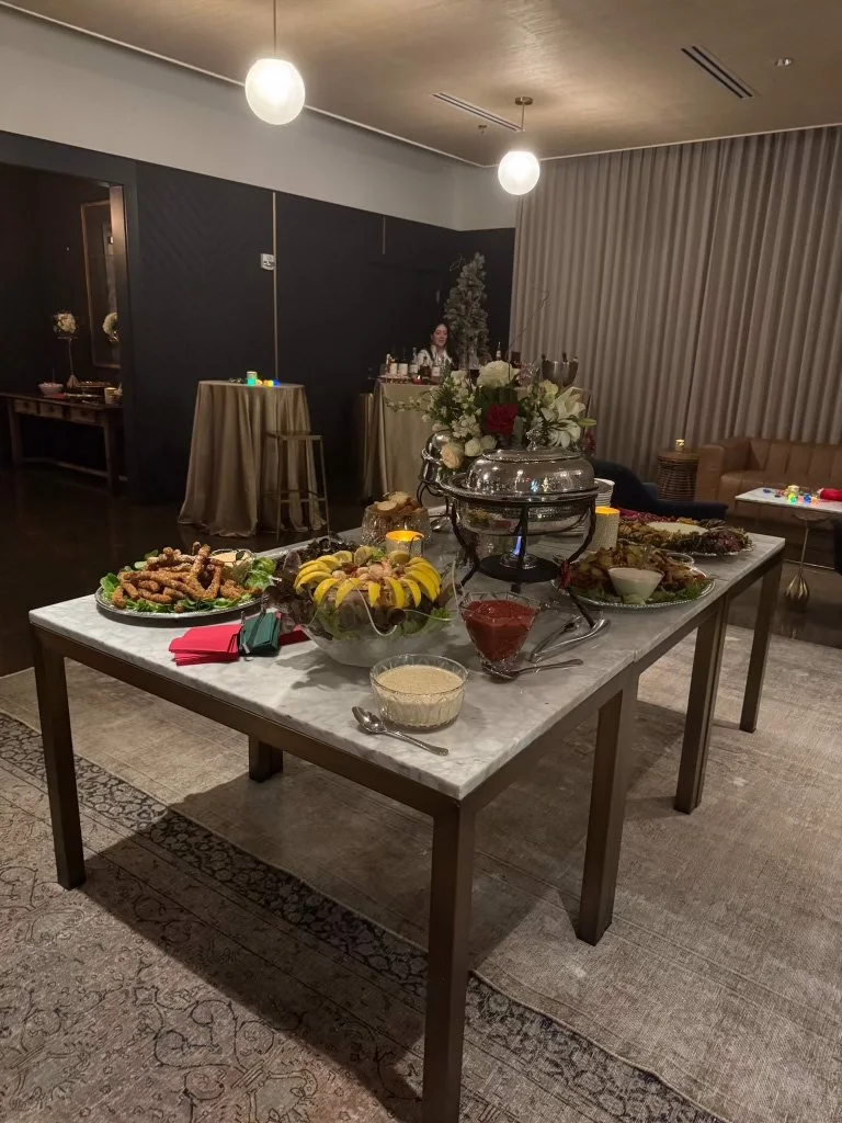 A table with various food items including a fruit platter, salads, and a hot dish, set up for a buffet or gathering in a modern room with dim lighting and minimal decoration.