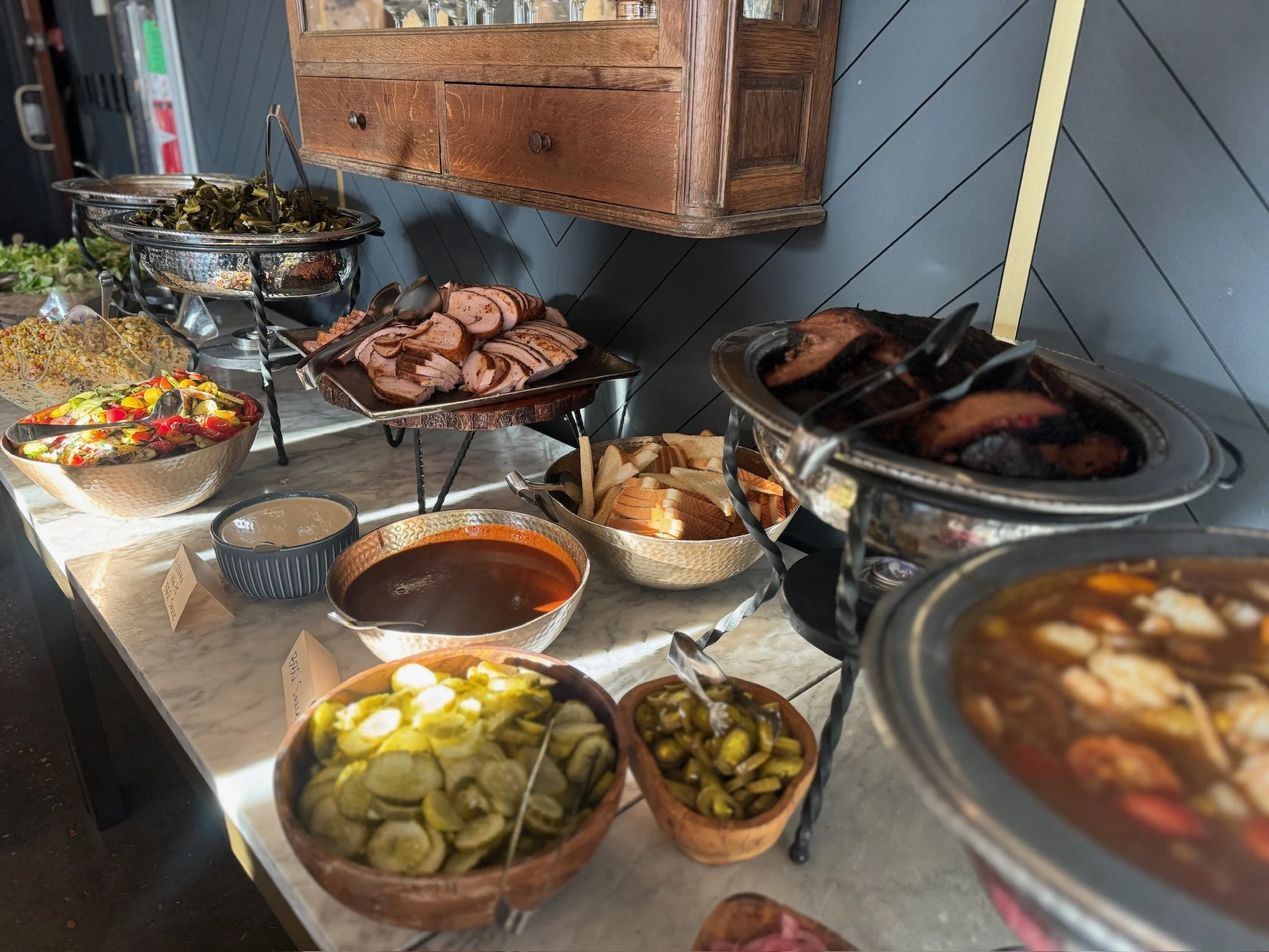 Buffet table with various dishes including sliced meats, salads, pickles, bread, and bowls of sauces and condiments.