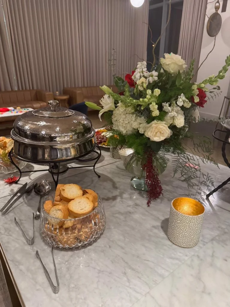 A marble table with a large floral arrangement, a covered dish, a glass bowl of sliced bread, and a decorative candle holder.