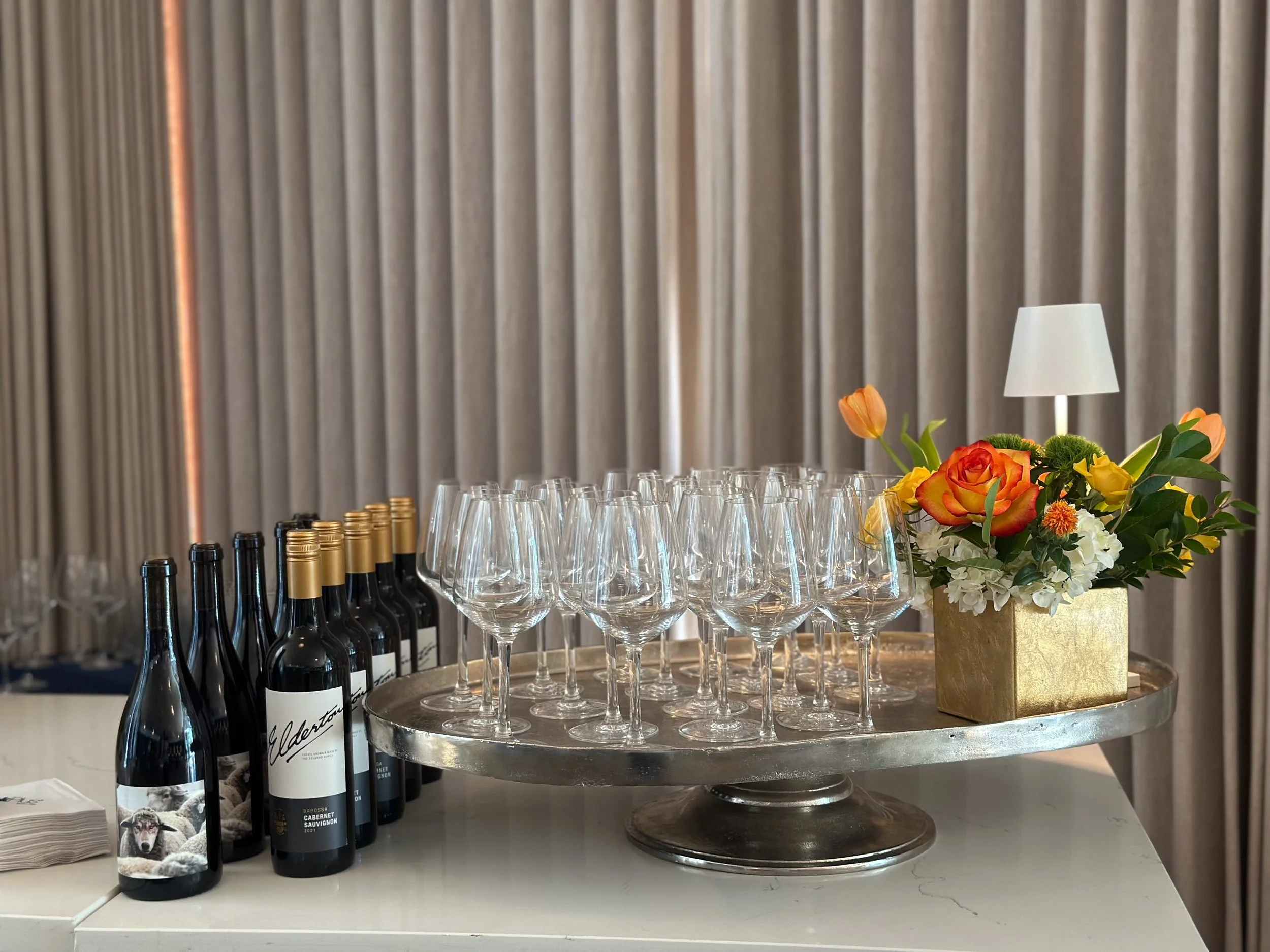 A table setup with wine bottles, empty wine glasses, and a floral arrangement in a gold container, with a small white lamp on a backdrop of gray curtains.