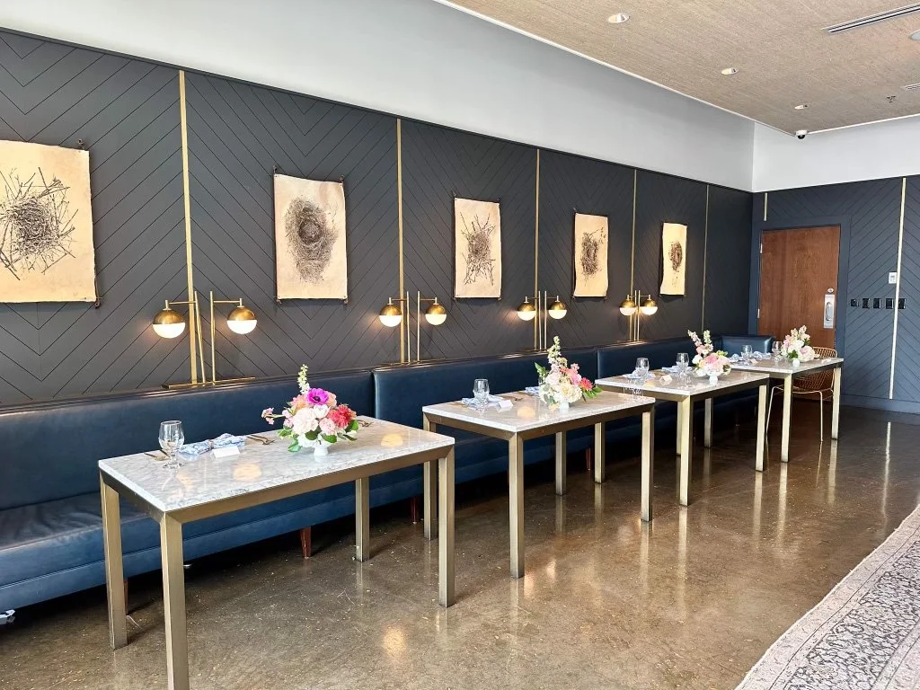 Empty dining tables with marble tops, floral centerpieces, wine glasses, and napkins in a modern restaurant with dark blue walls, framed abstract artwork, black leather seating, and gold lighting fixtures.