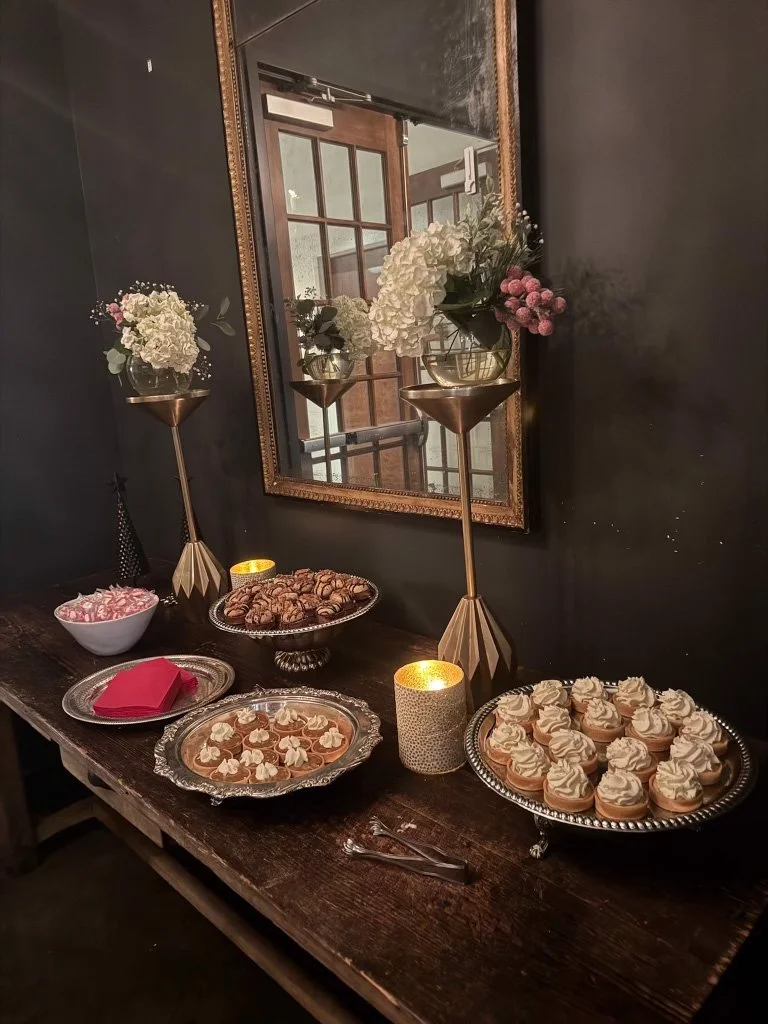 A dessert table with cupcakes, cookies, and candies, lit by candles and decorated with flowers, mirror, and candle holders in an elegant setting.