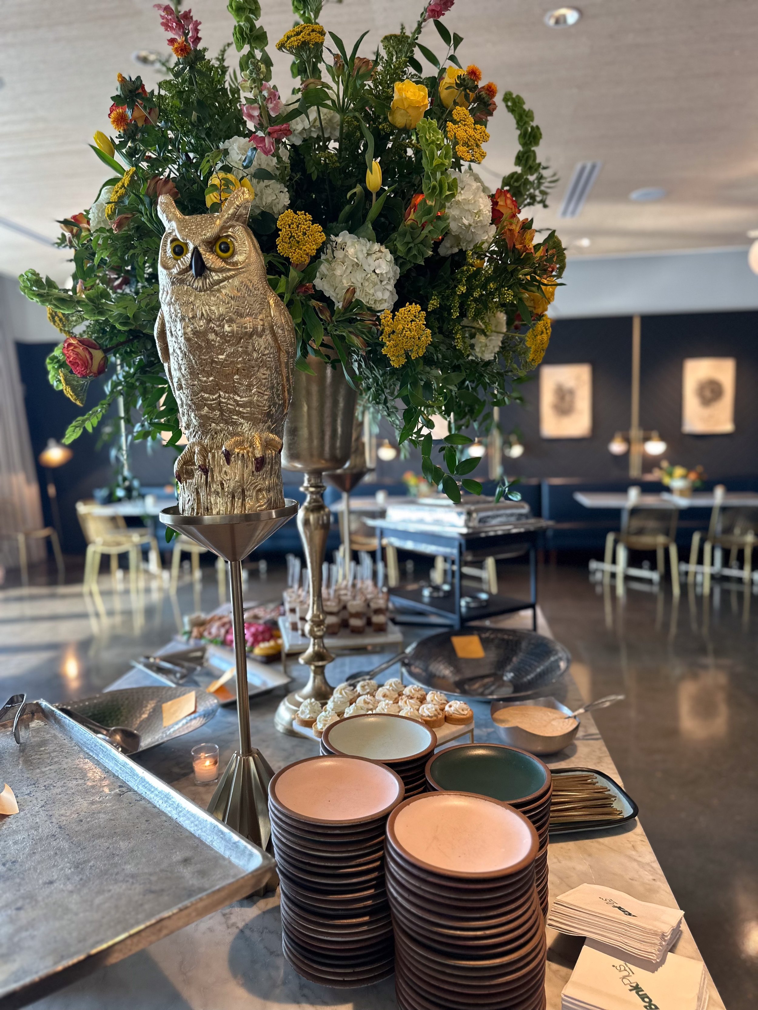 A decorative golden owl figurine on a stand in front of a large floral arrangement on a table, with plates and utensils while inside a dining area.