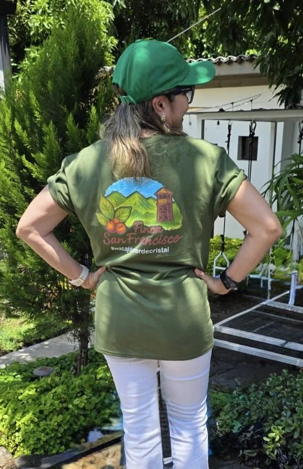 Back view of a woman wearing a green baseball cap, glasses, a green shirt with a colorful embroidered patch of a landscape, and white pants, standing outdoors with greenery and a white building in the background.