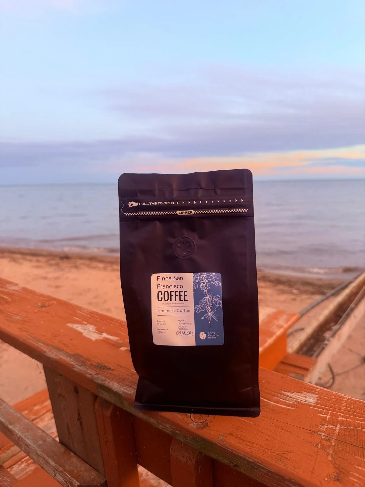 A black bag of coffee labeled 'Finca San Francisco Coffee' and 'Pacamara Coffee' with a floral illustration, placed on a wooden railing along a beach or lakeside, with water and a cloudy sky in the background during sunset.