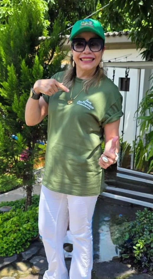 Woman smiling outdoors, wearing sunglasses, a green cap, green t-shirt, and white pants, pointing at her shirt.