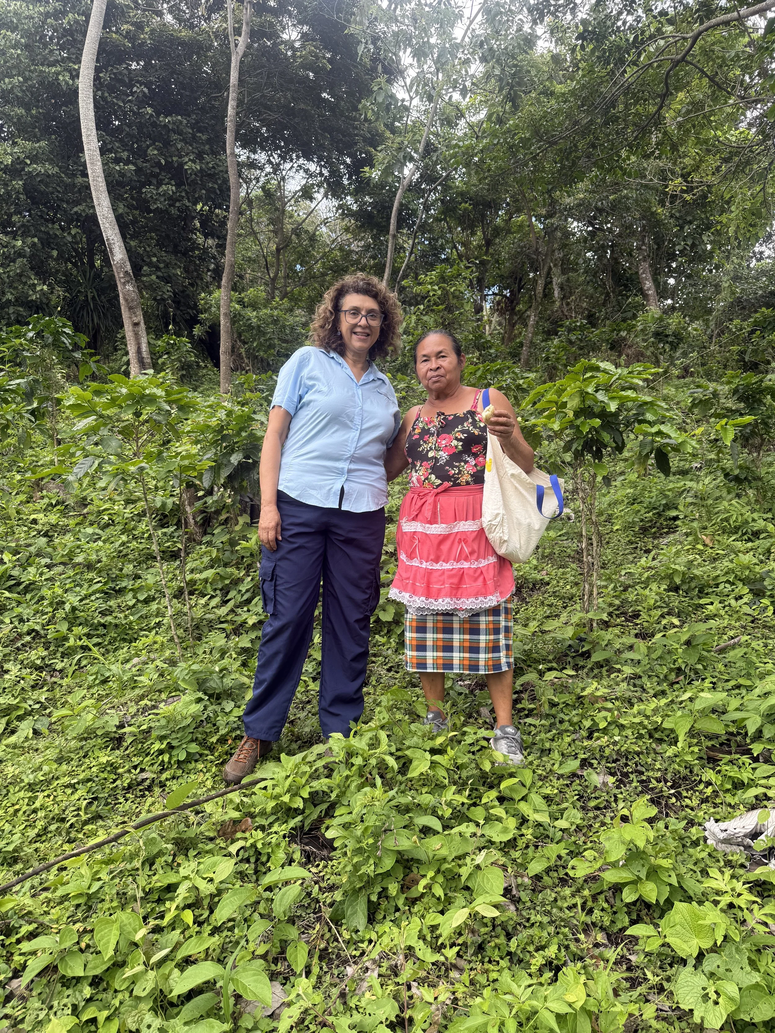 Two women standing in a lush green forest with trees and leafy plants in the background. One woman is wearing a light blue shirt and navy pants, and the other is in a floral top with a pink skirt and a plaid skirt underneath. They are smiling and posing for the photo.