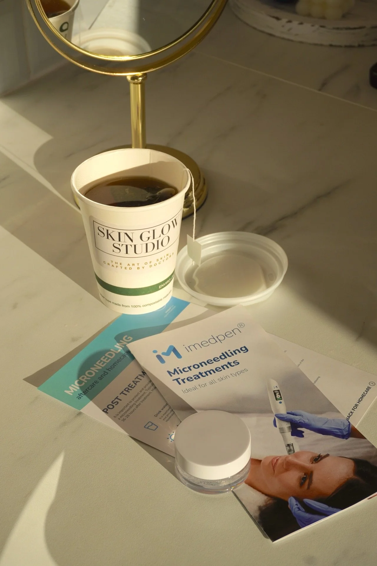 A cup of black coffee on a vanity table beside a small mirror, skincare brochures, a small jar, and a box of micro-needling treatments.