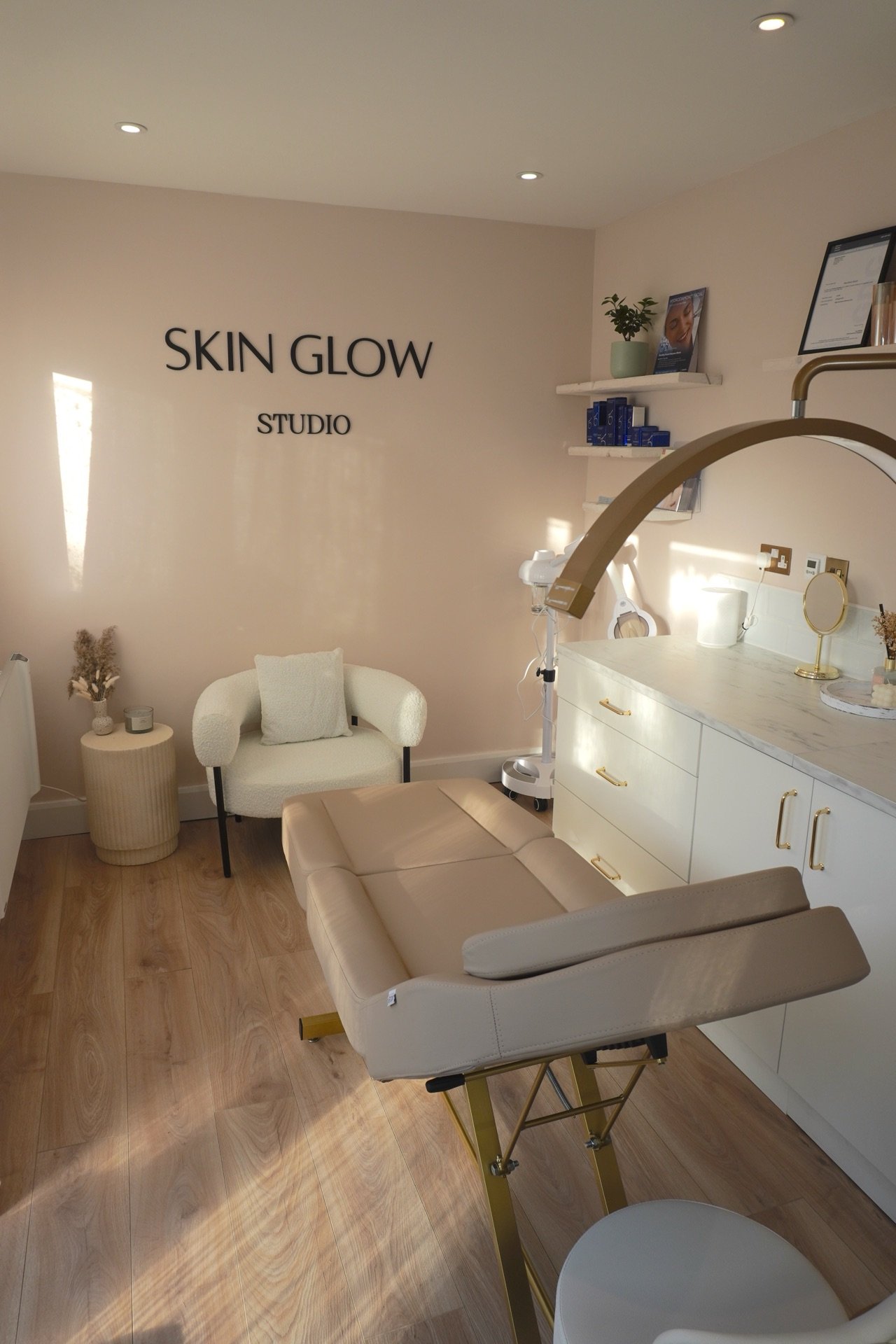 Interior of a skincare studio with a treatment bed, white cabinetry, and shelves with skincare products, decorated with plants and framed photos.