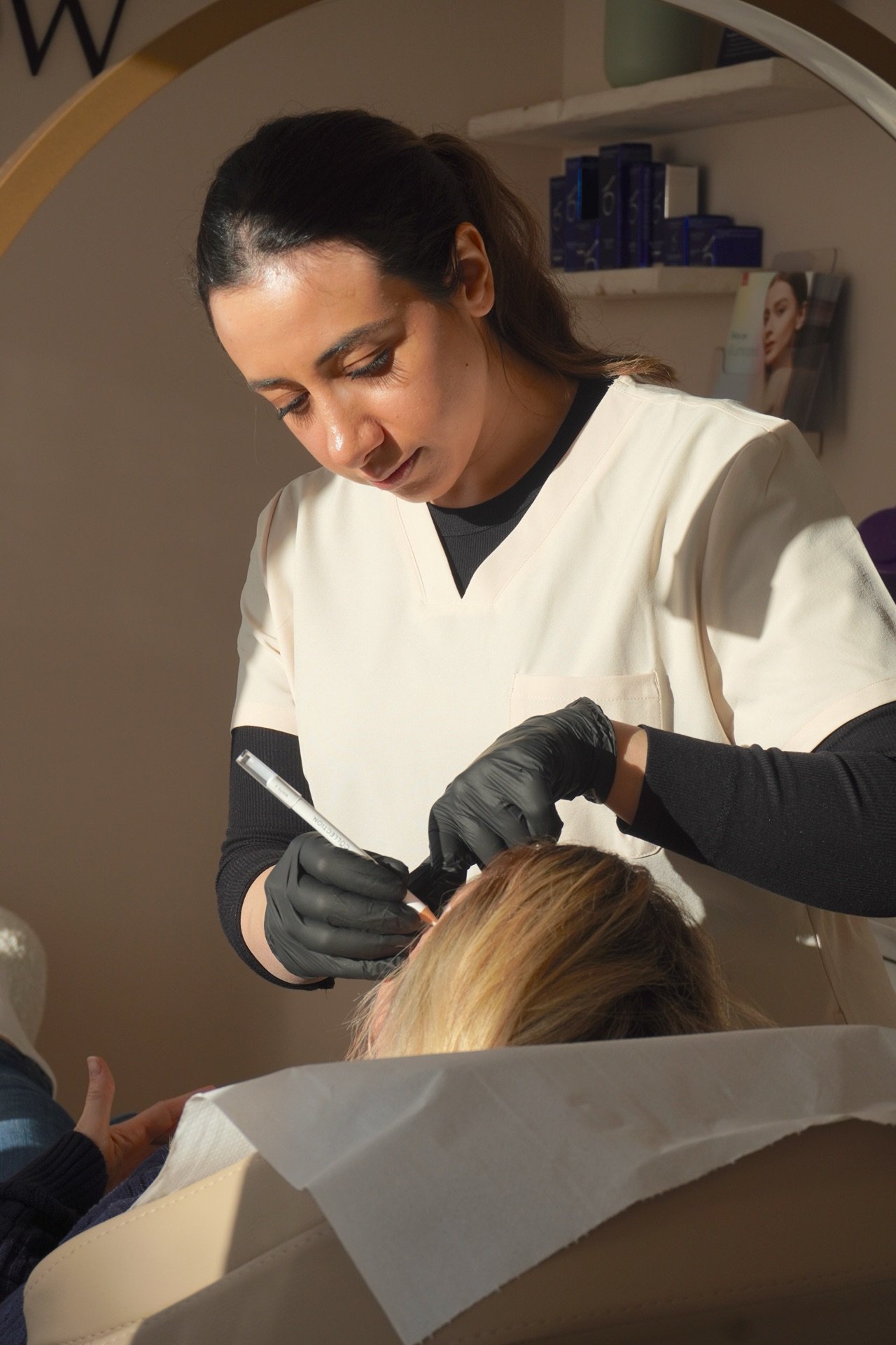 A female tattoo artist in a white uniform and black gloves is tattooing a person's forehead in a professional studio setting.