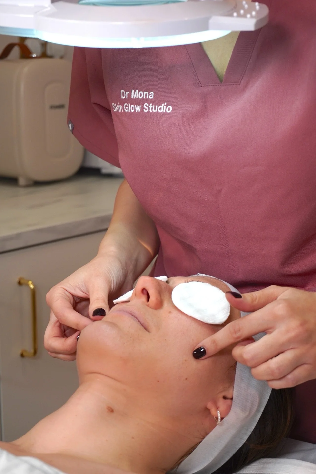 A woman is receiving a facial treatment at a skincare studio, with her eyes masked and a specialist attending to her face.