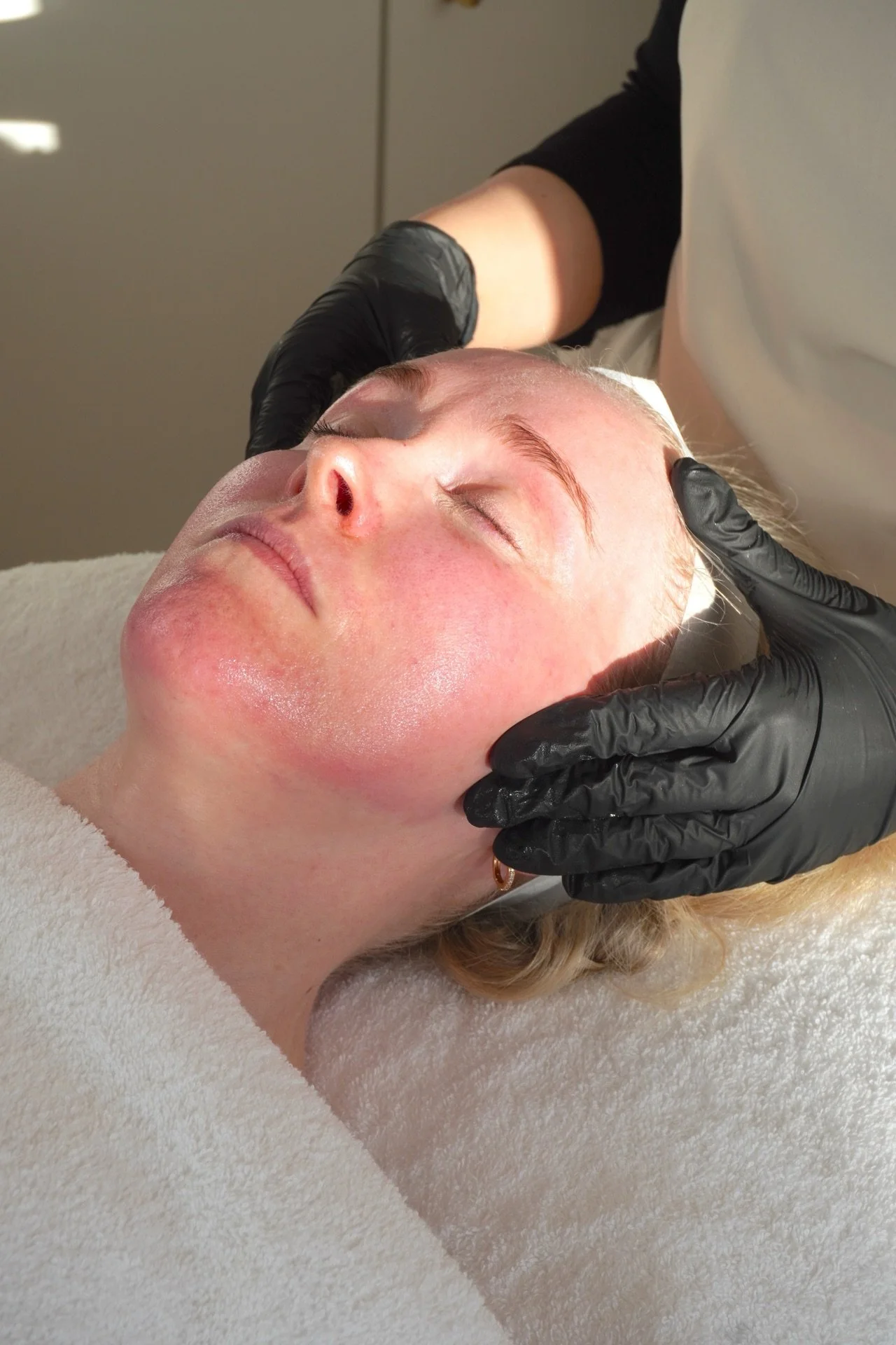 A woman receiving a facial treatment at a spa, lying down with eyes closed, while a technician in black gloves gently touches her face.