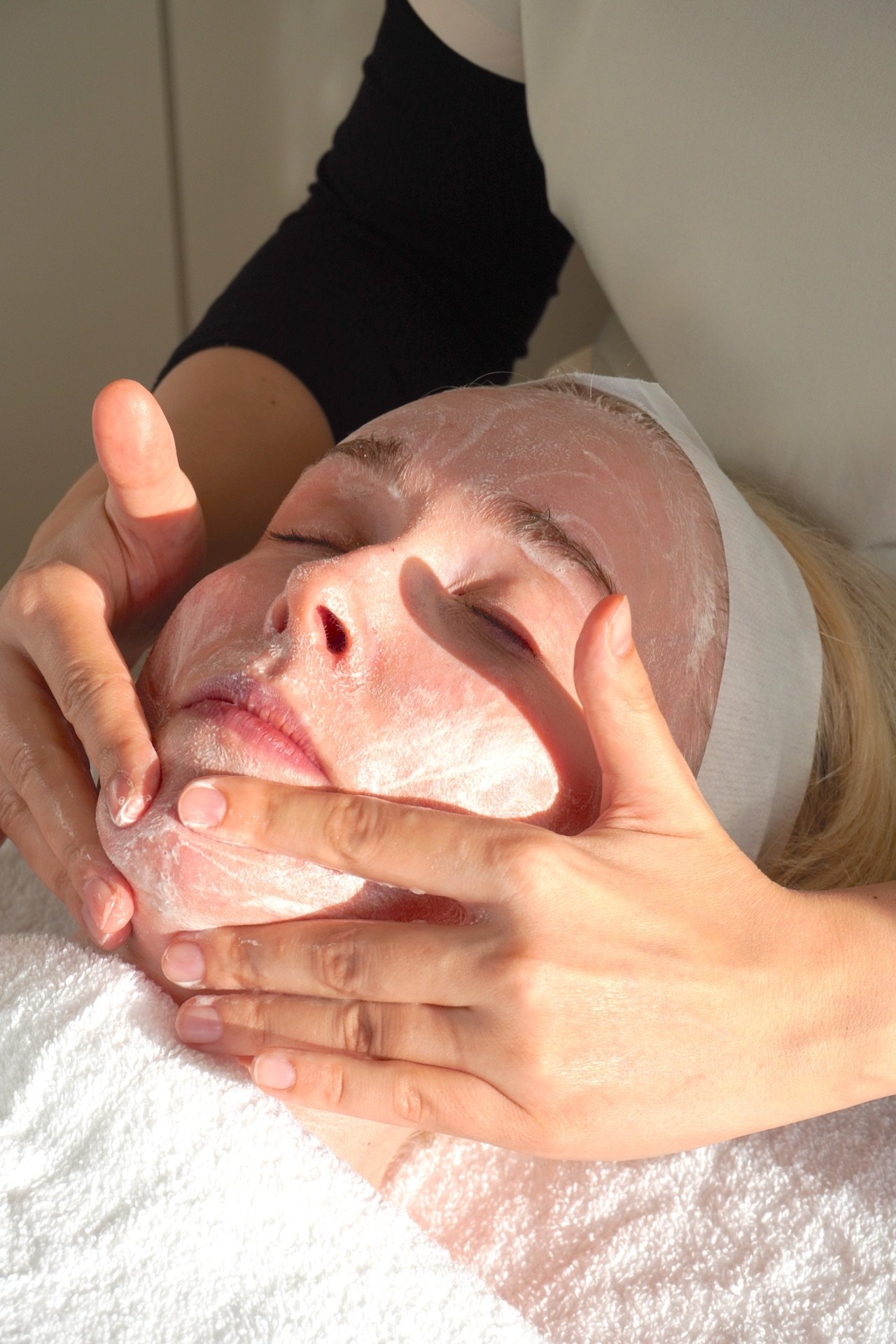 A woman receiving a facial treatment, lying down with her eyes closed, while a beautician applies a facial mask or cream.