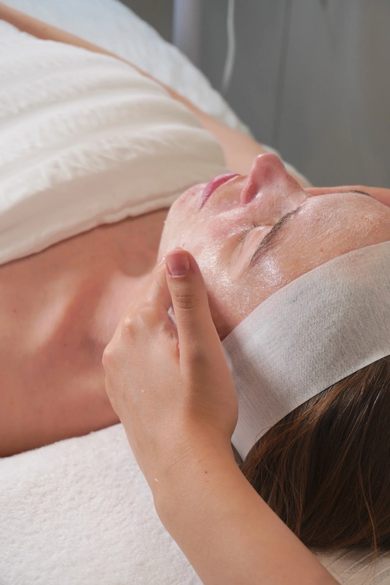A woman lying on a massage table during a facial treatment, with a towel across her chest and a headband covering her hair, receiving a facial massage from a therapist.