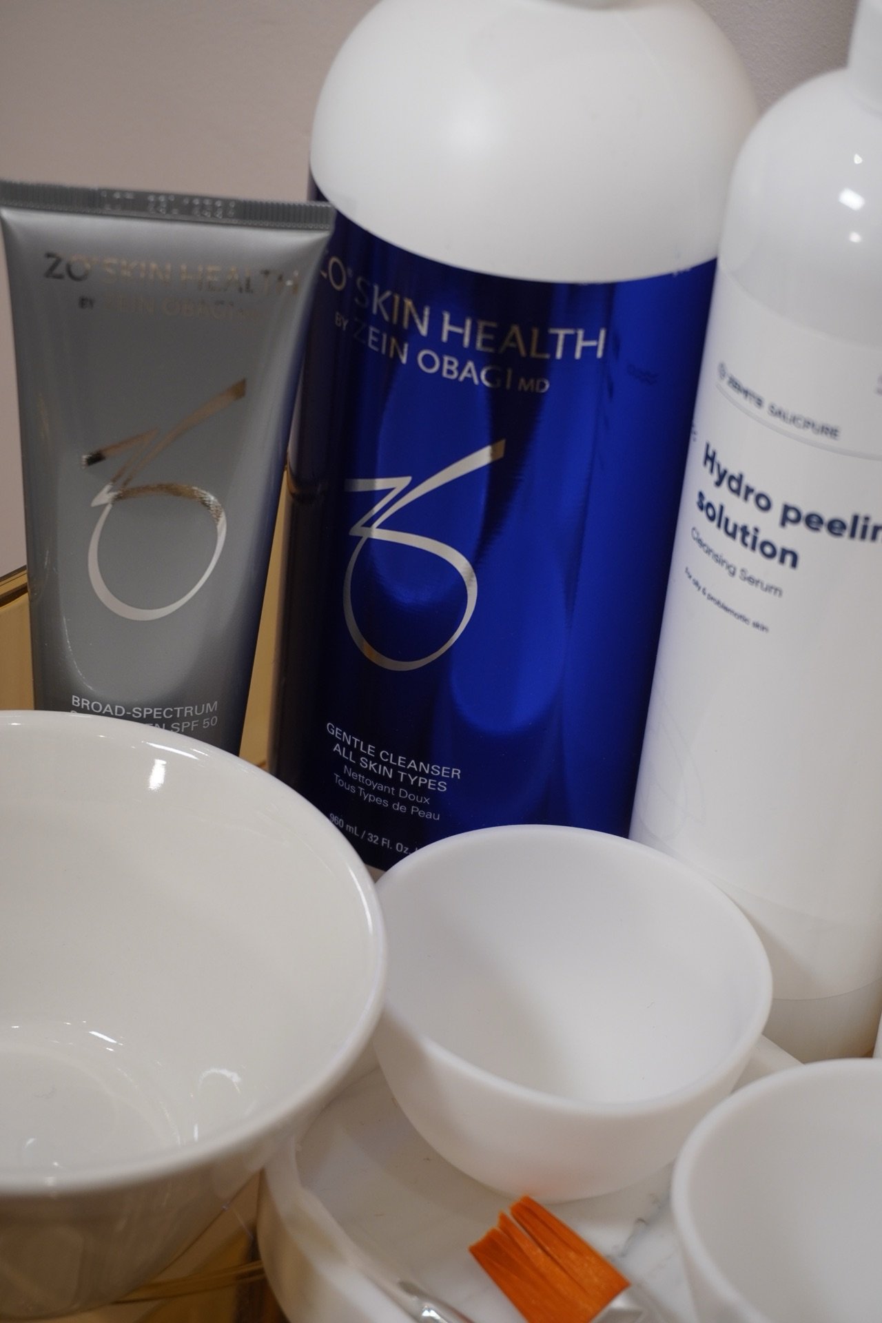 Skincare products and white bowls on a surface, including a ZO Skin Health Broad Spectrum SPF 50, a blue bottle of ZO Skin Health Gentle Cleanser, and a white bottle of Hydro Peeling Solution.