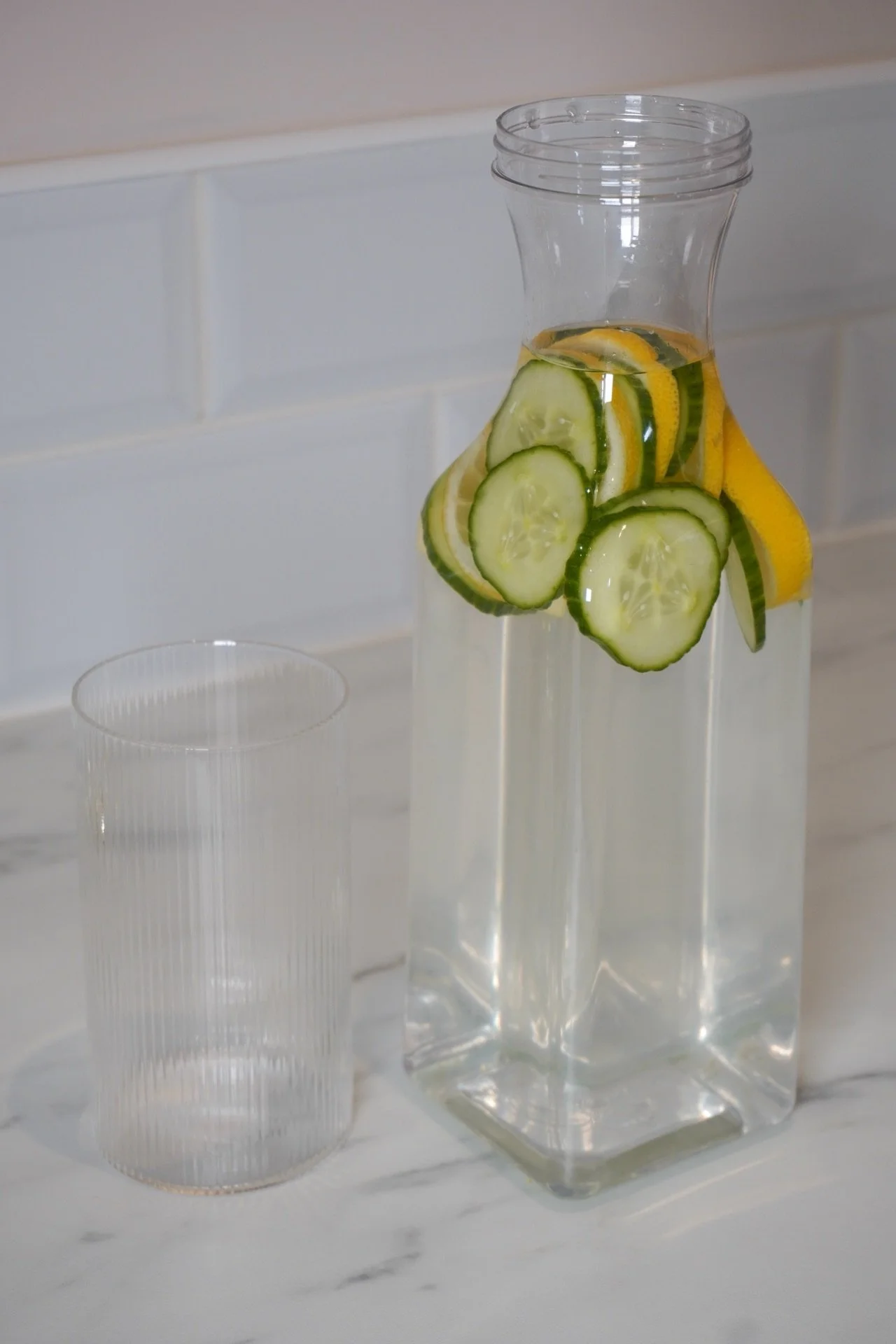 A glass pitcher filled with water and slices of cucumber and lemon, standing next to a clear glass tumbler on a white marble surface.