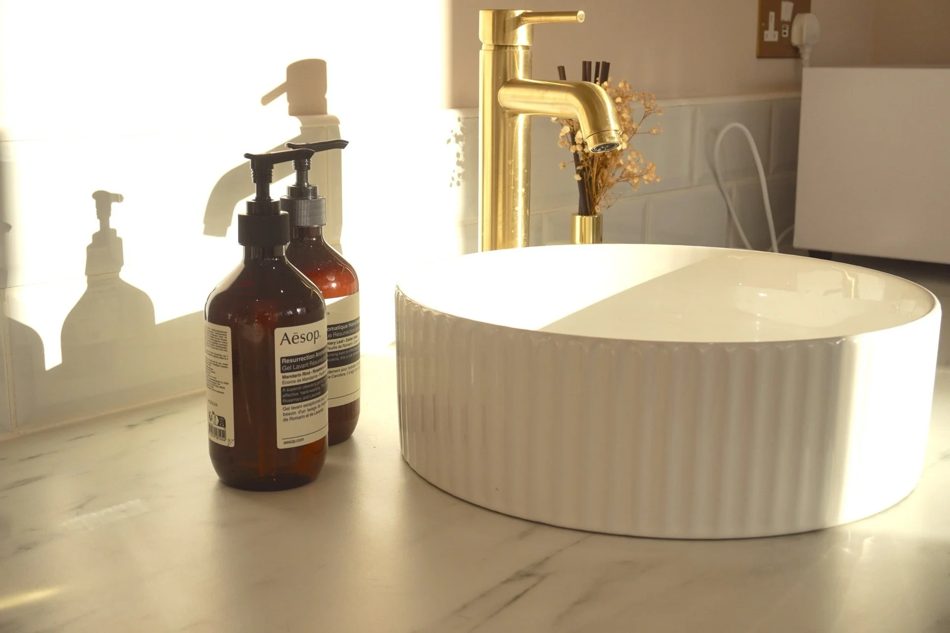 A white round basin sink with vertical ridges and a gold faucet, two amber hand soap bottles labeled Aesop, and a small vase with dried flowers on a marble countertop, illuminated by natural light creating shadows on the wall behind.