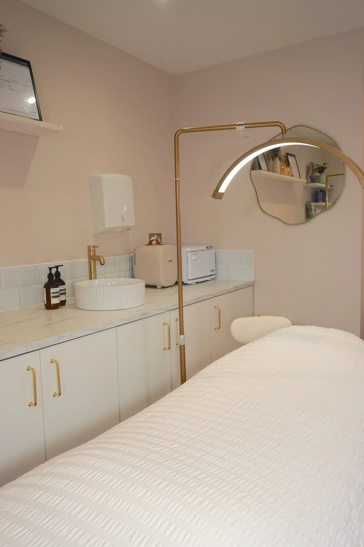 A clean and modern spa treatment room with a massage table covered in a white textured blanket, a circular sink with a gold faucet, bottles of soap, a small microwave, and a mirror reflecting shelves with decor and skincare products.