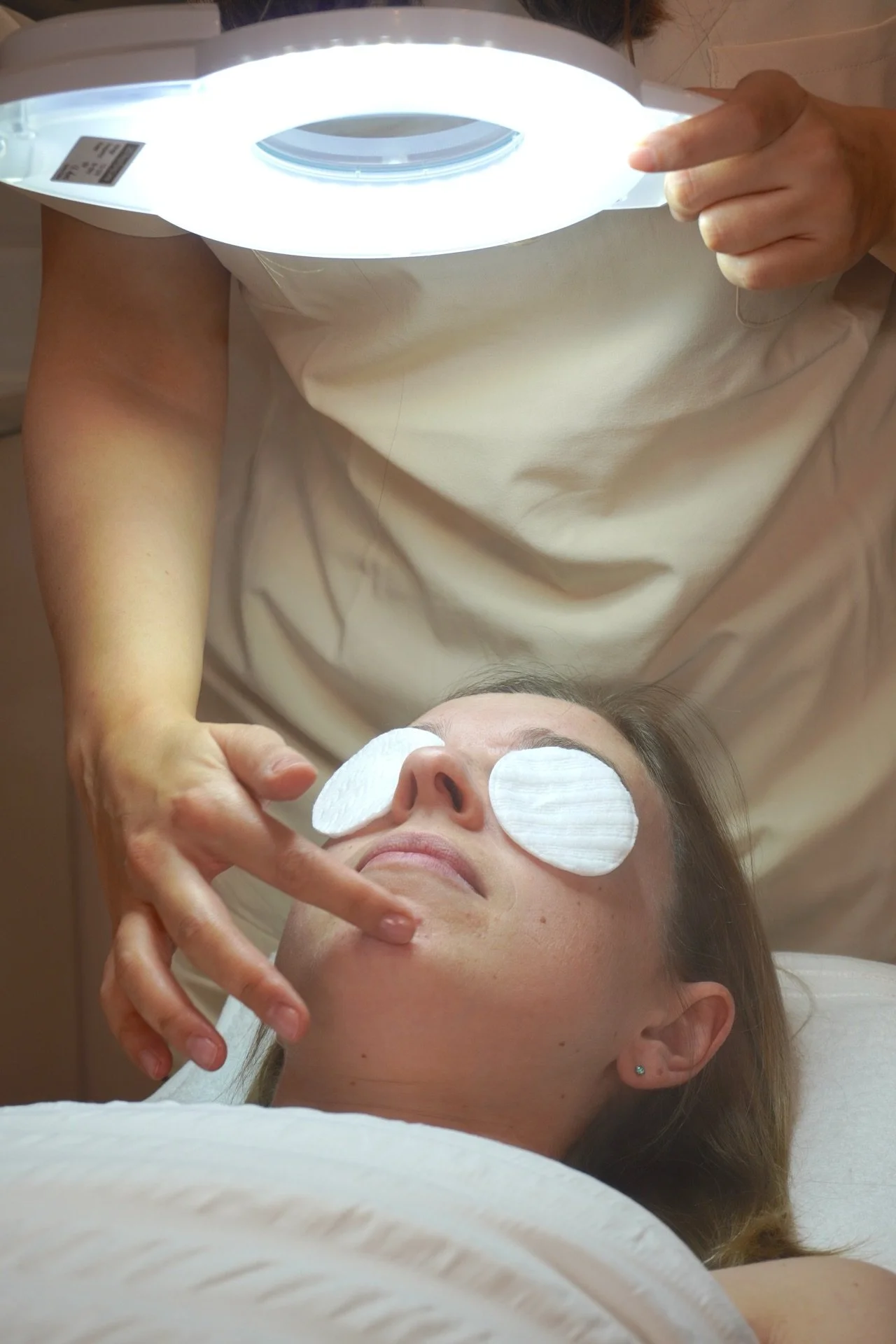 A woman receives a facial treatment with white eye patches and cotton pads on her face, lying on a treatment bed while a beauty professional holds a lamp above her face.
