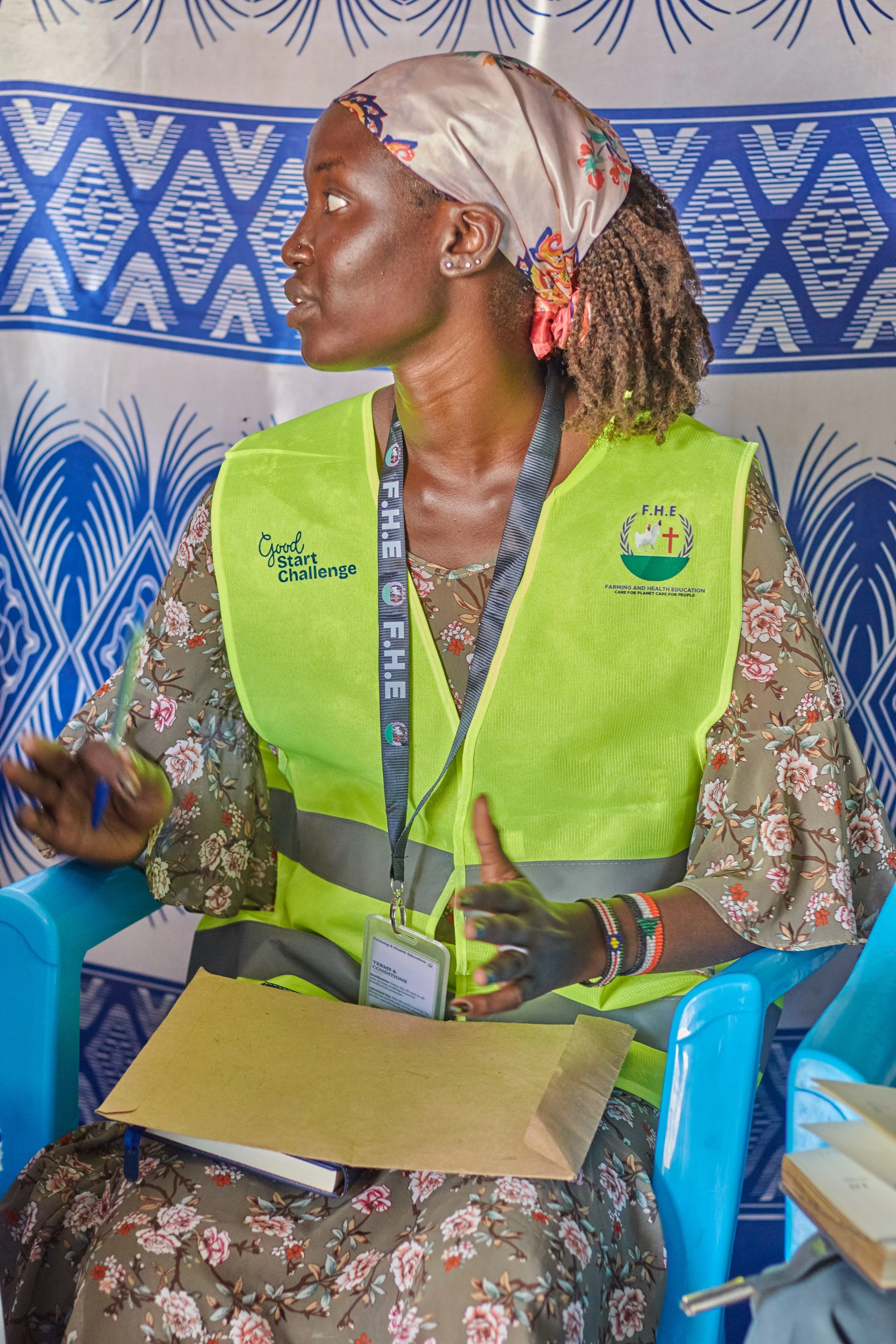 A woman wearing a neon green vest with the words 'Good Start Challenge' and a logo that says 'Farming and Health Education' is seated at a table. She has a floral patterned shirt underneath and a colorful headscarf. She is wearing earrings, a lanyard