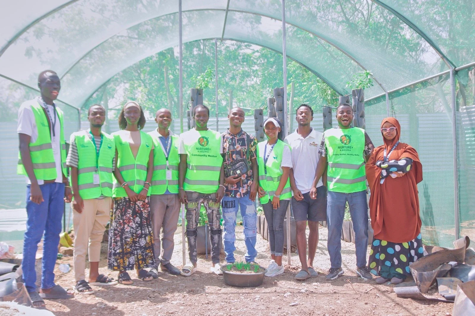 A group of eleven people standing together inside a greenhouse or plant nursery. Some are wearing green vests and others are in casual or traditional clothing. The individuals are smiling and posing for the photo, with small plants visible in the for