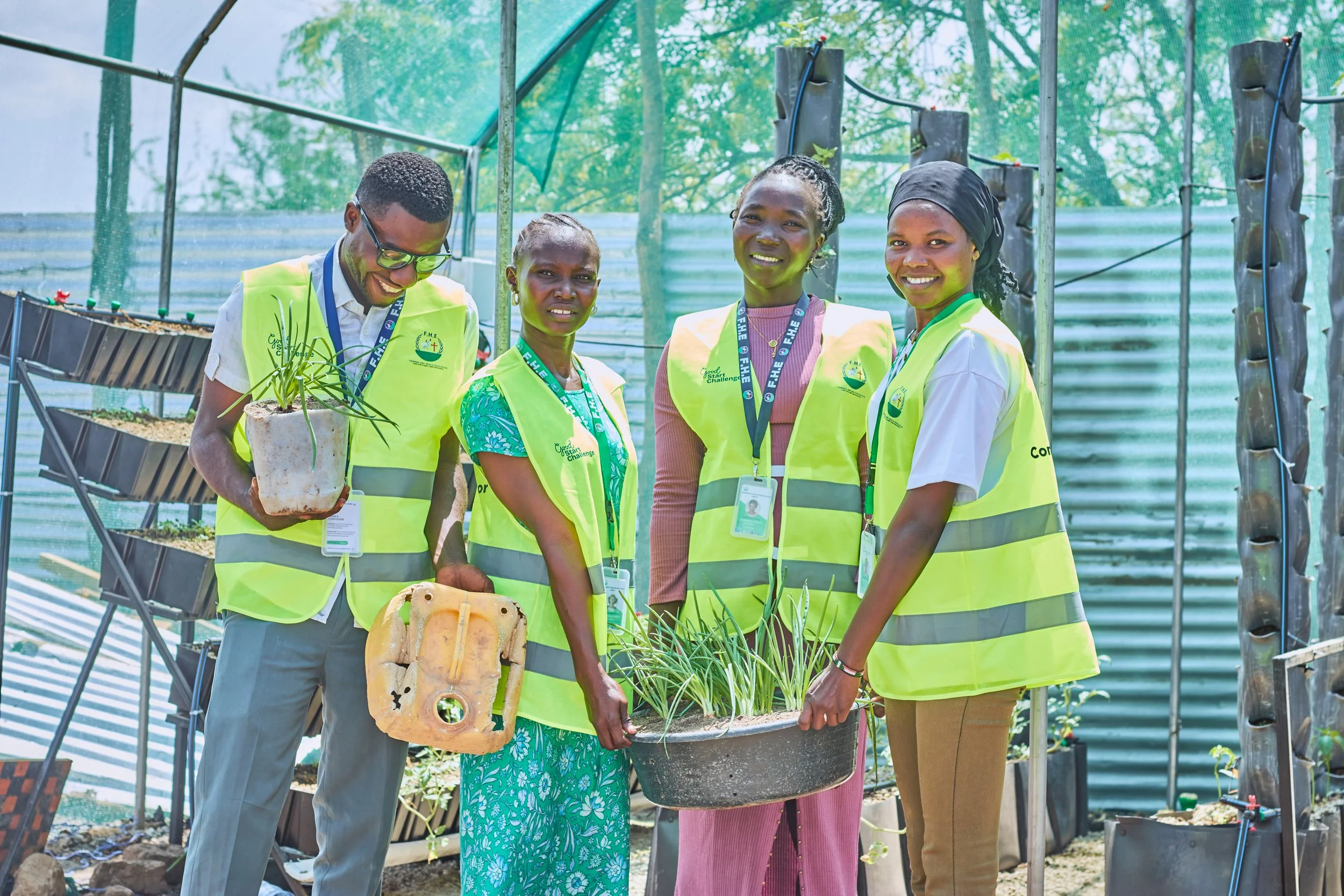 Group of four people standing in a greenhouse, holding plants and gardening tools, wearing yellow safety vests with logos, smiling at the camera.