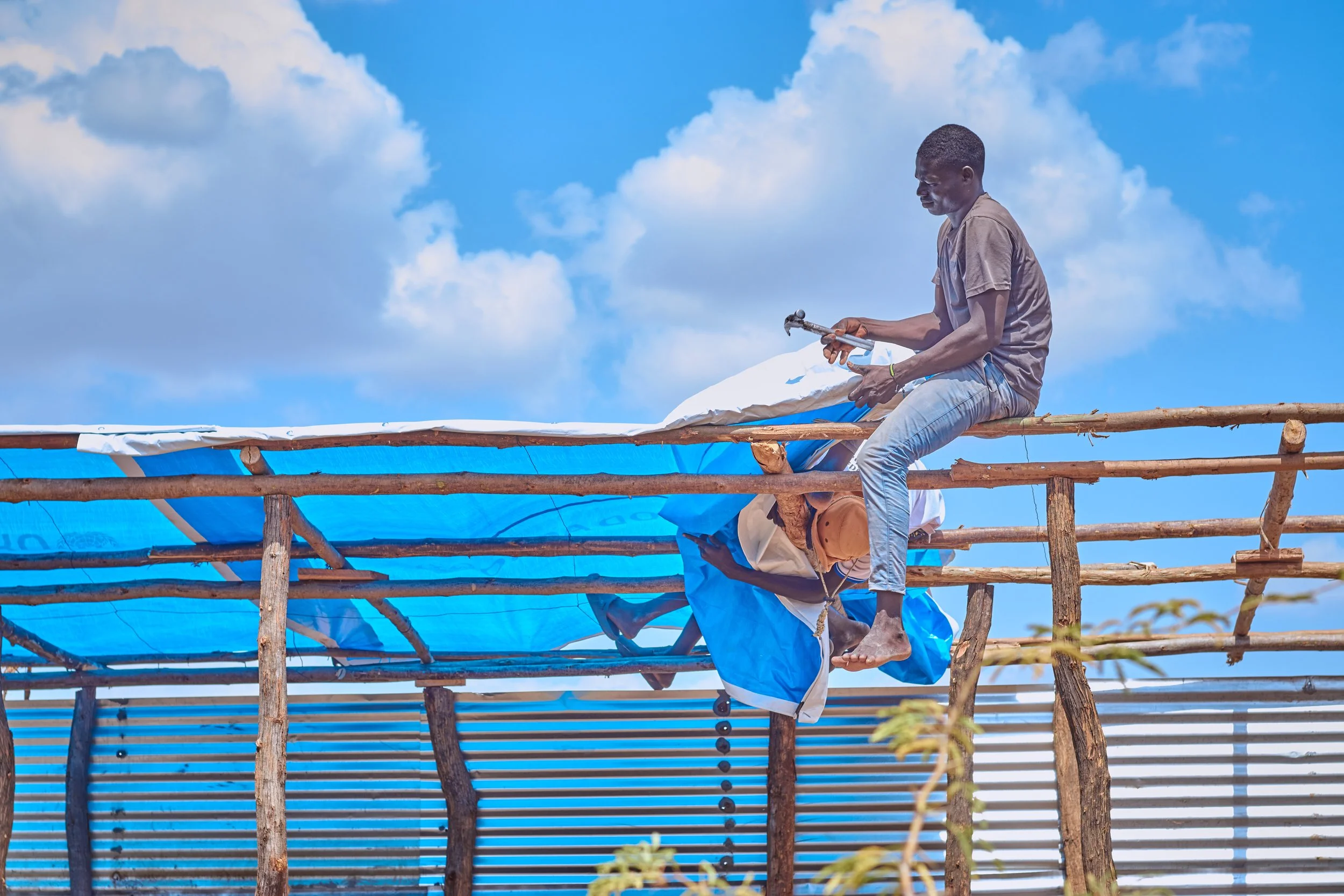Two men working on a bamboo structure under a bright blue sky with clouds. One man is sitting on the bamboo beams, and the other is holding a hammer, both engaging in construction work.