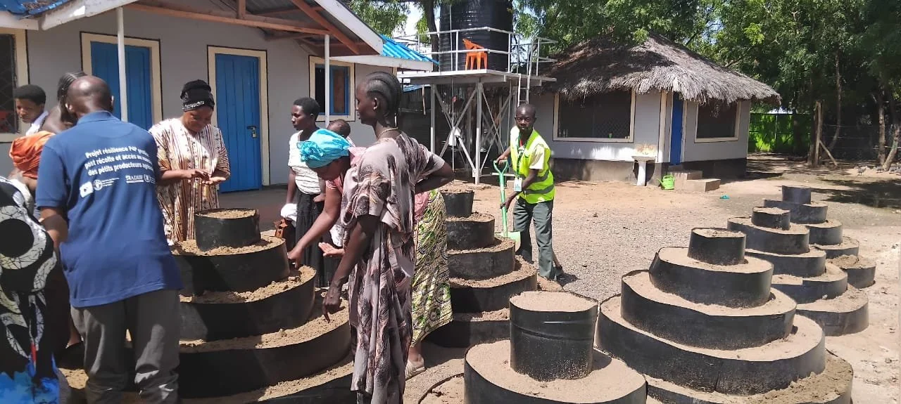 Group of people working together to build a traditional underground stove with round stages in a rural area, with houses and trees in the background.
