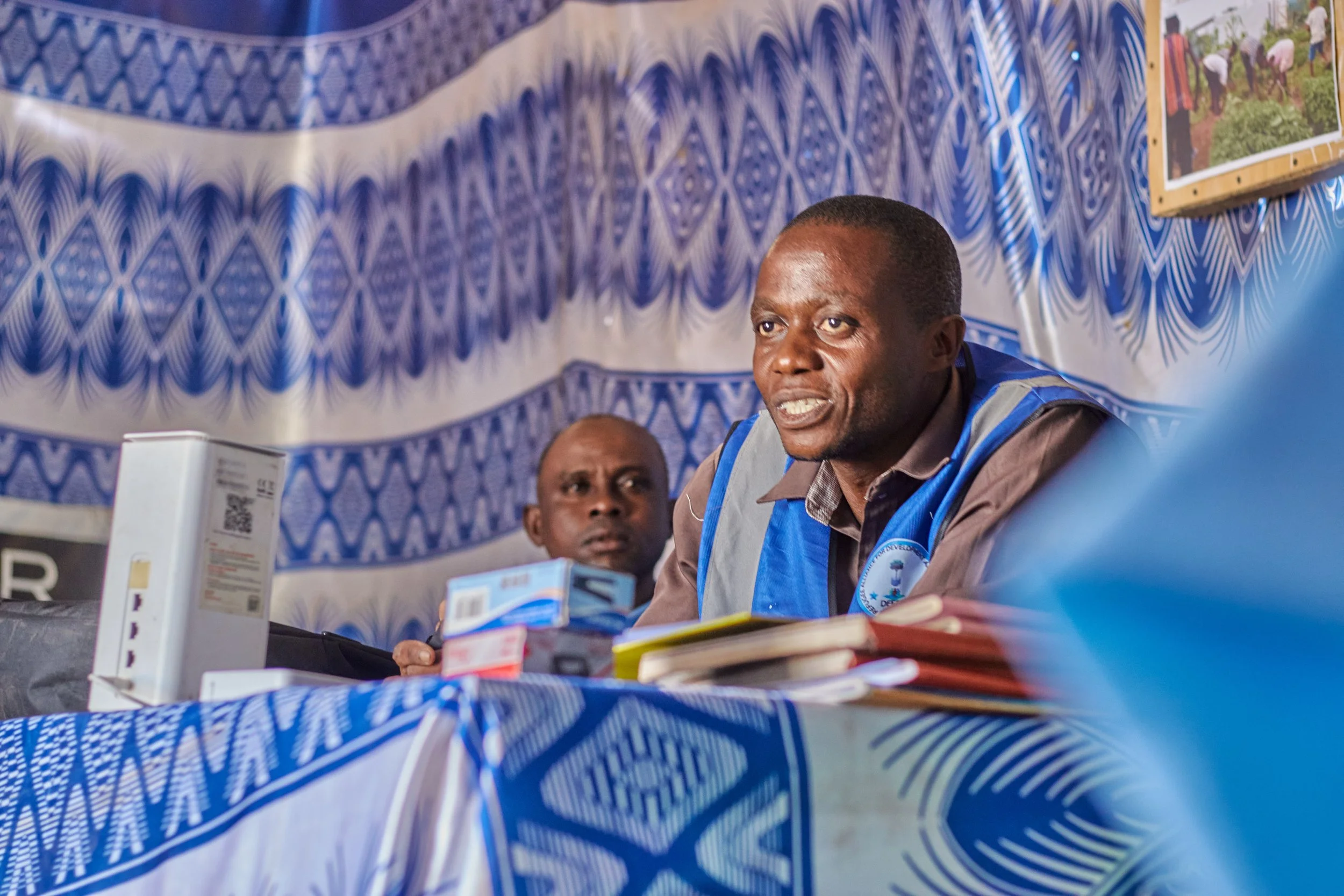 Two men in blue vests sitting at a table with various documents and medication bottles in front of them, inside a room with blue and white patterned walls and a framed photograph on the wall.