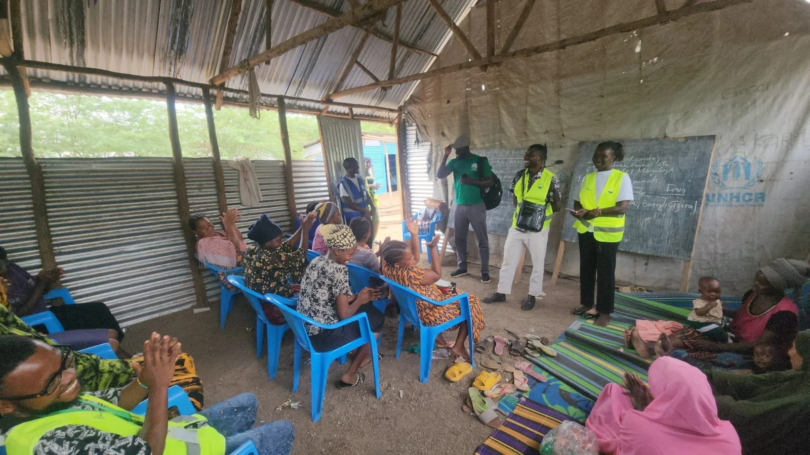 A community meeting held inside a rustic, corrugated metal-walled building. Several people, including women and children, are sitting on blue and striped mats and chairs. Some women are raising their hands, possibly asking questions or participating.