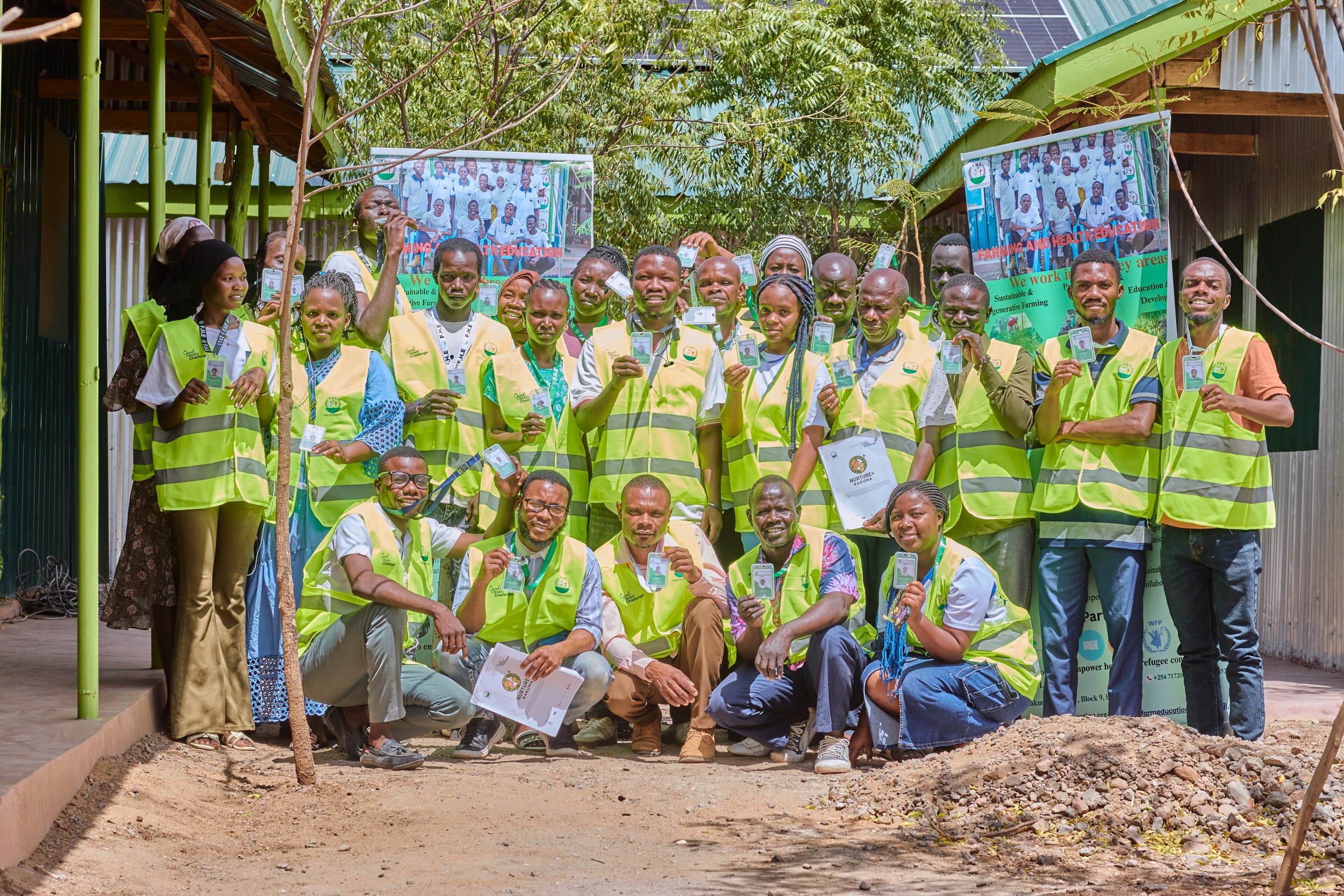 Group of people wearing yellow safety vests, holding identification cards, standing outdoors in front of a green building with trees, some are kneeling in front while others stand behind, with two posters in the background promoting health and agriculture education.