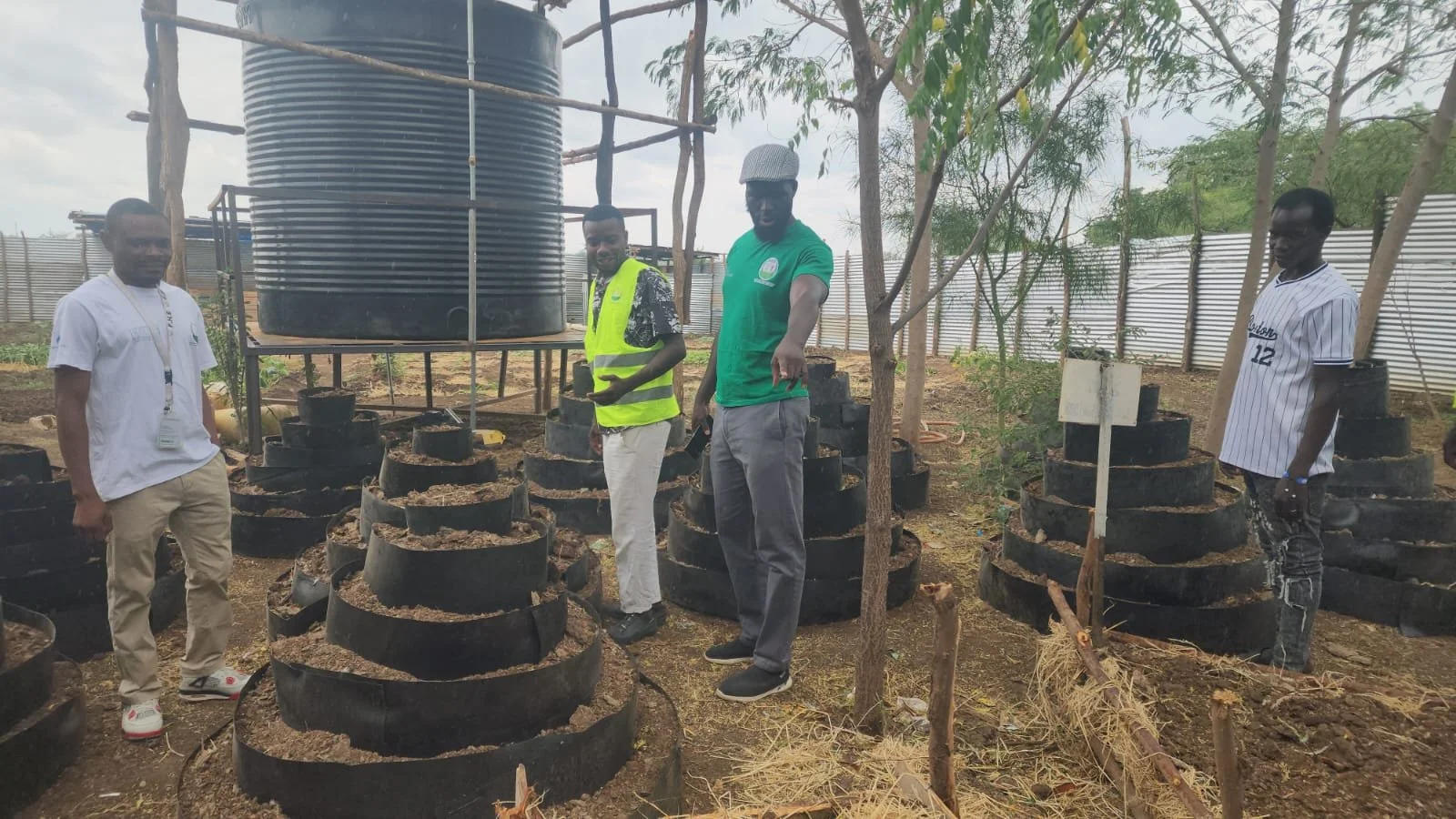 Four men standing in a garden with a large water tank in the background. They are surrounded by black plastic plant containers filled with soil. The men are dressed casually, and some are looking at the camera. There are small trees and a white fence