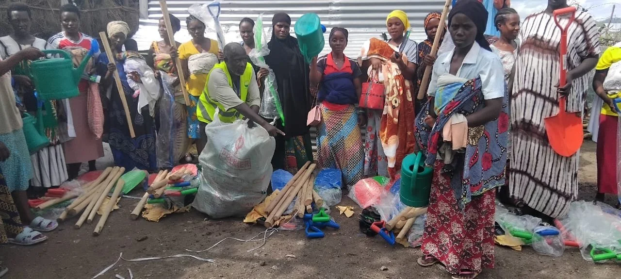Group of women wearing colorful clothing and headscarves standing outdoors with gardening and farming tools like watering cans, shovels, and hoes displayed on the ground.