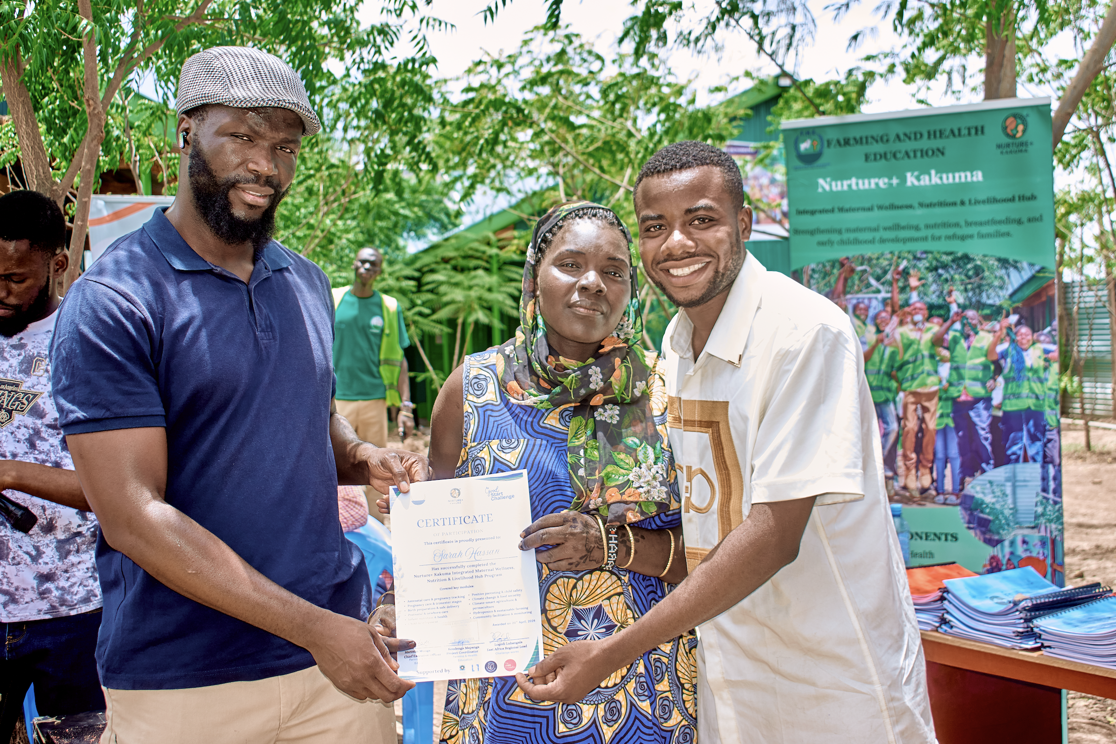 Group of people at an outdoor event, with a woman receiving a certificate from a man, surrounded by trees and a banner about farming and health education.