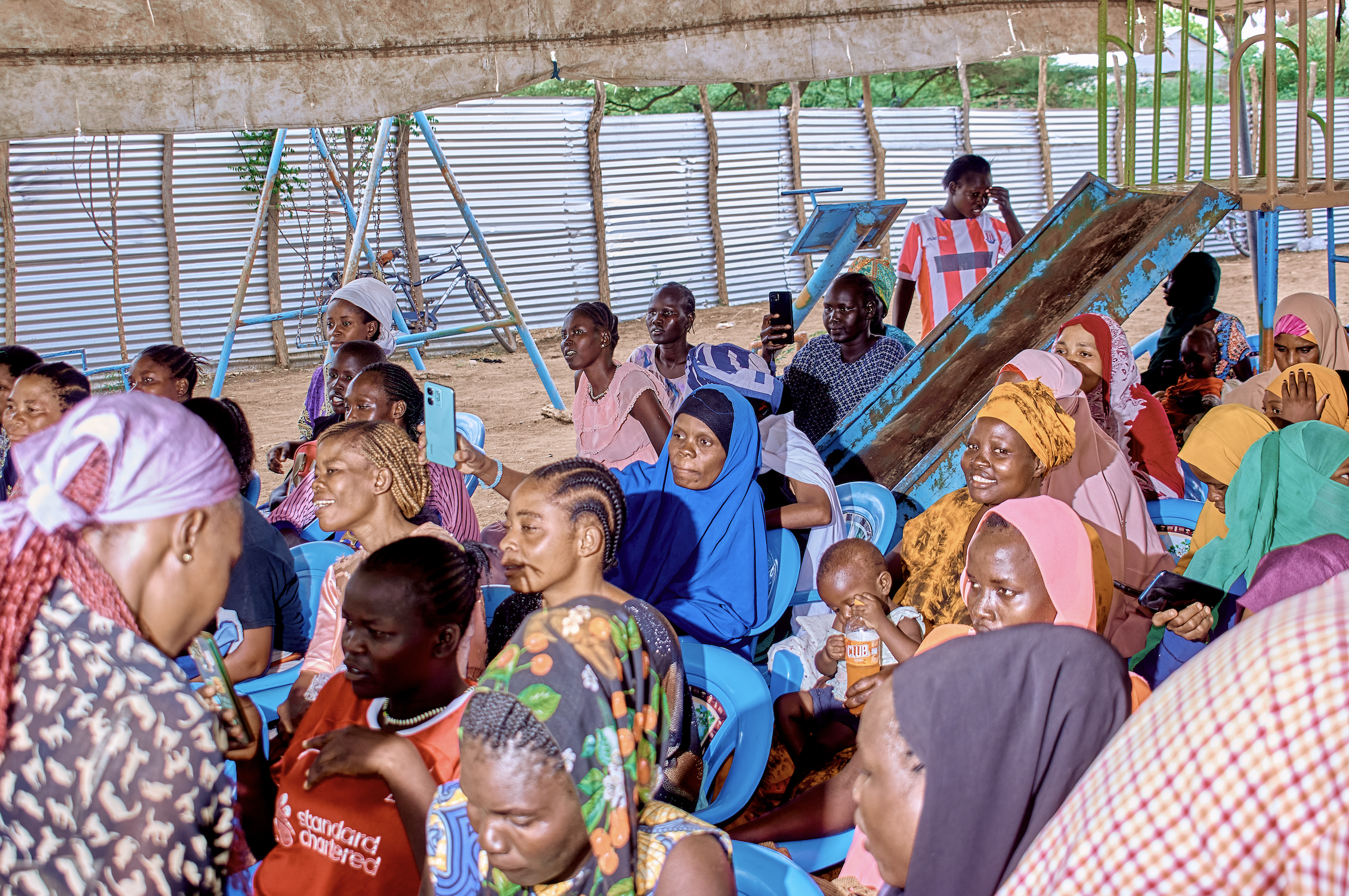A group of women and children gathered under a makeshift shelter with a corrugated metal wall behind them. Some are sitting on blue plastic chairs, others standing. Many are holding or looking at their phones. There are swings and a slide in the back
