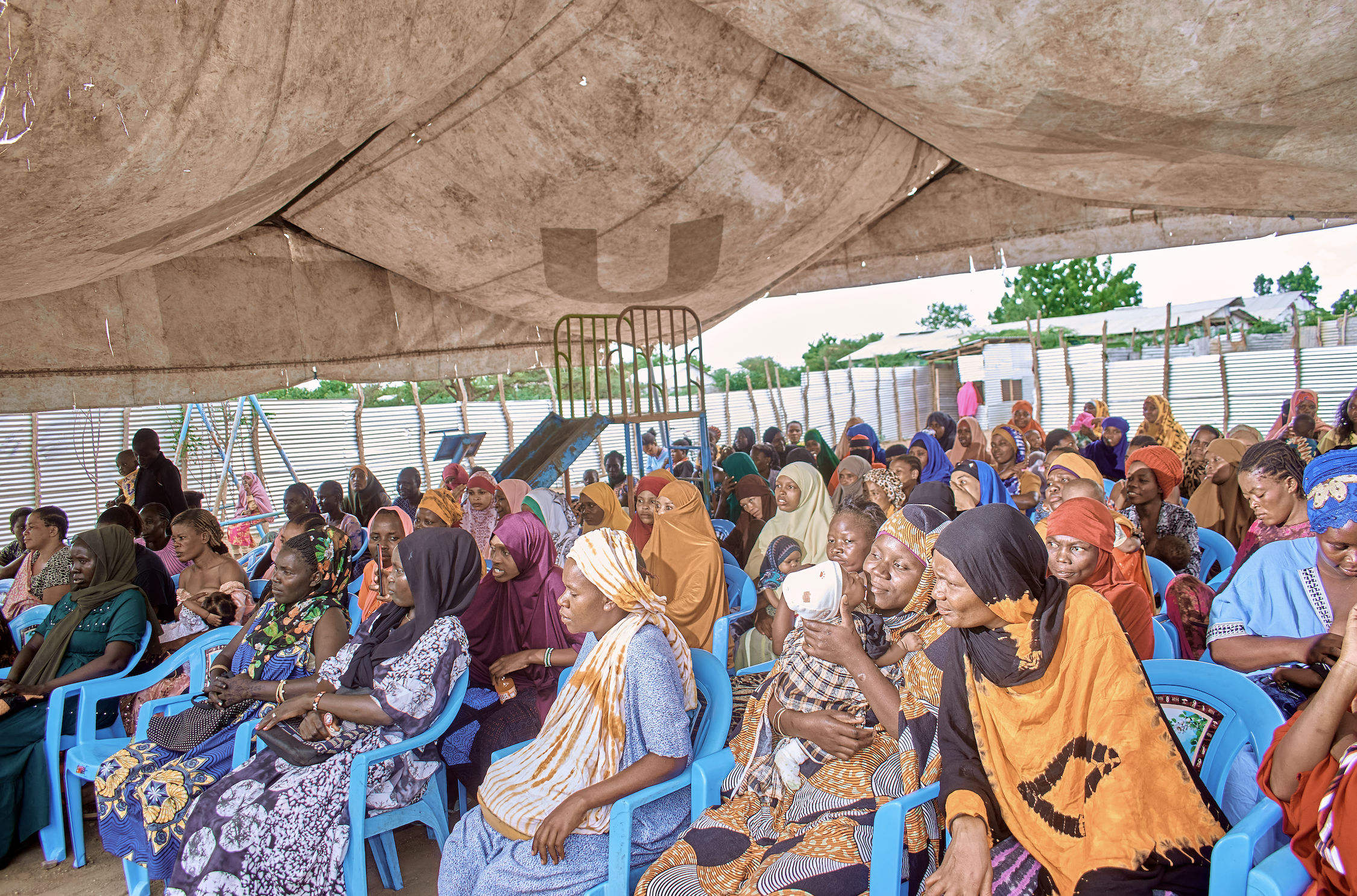 A large gathering of diverse women and children seated in blue plastic chairs outdoors beneath a makeshift shelter made of fabric, with trees and simple structures visible in the background.