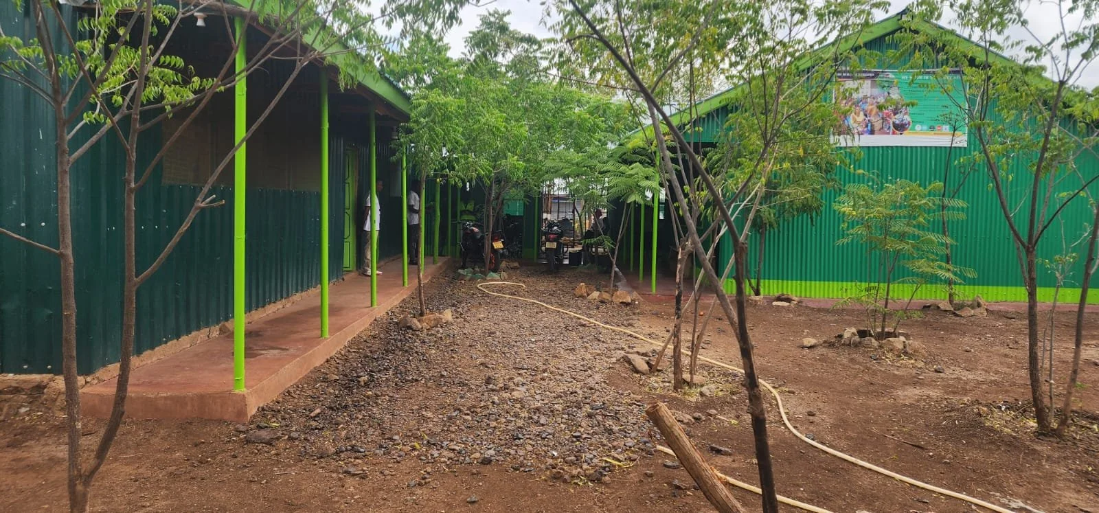 A green metal structure with lime green support poles and a pathway in the middle, surrounded by trees and soil, with a few motorcycles parked under the shade of trees in the background.