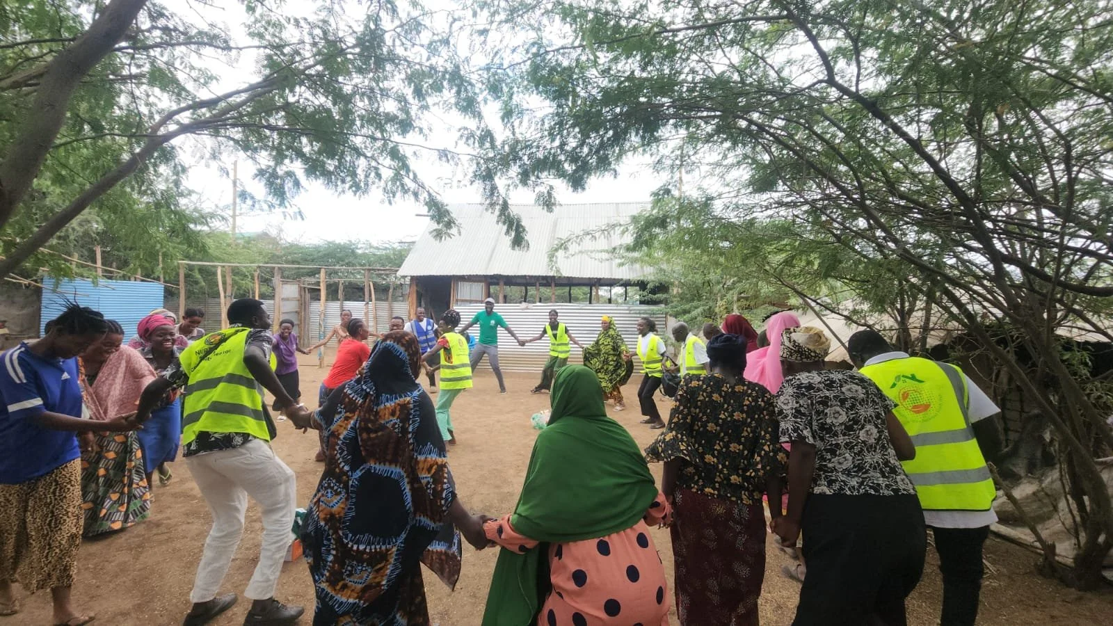 Group of people holding hands and dancing in an outdoor area surrounded by trees, with a building in the background.