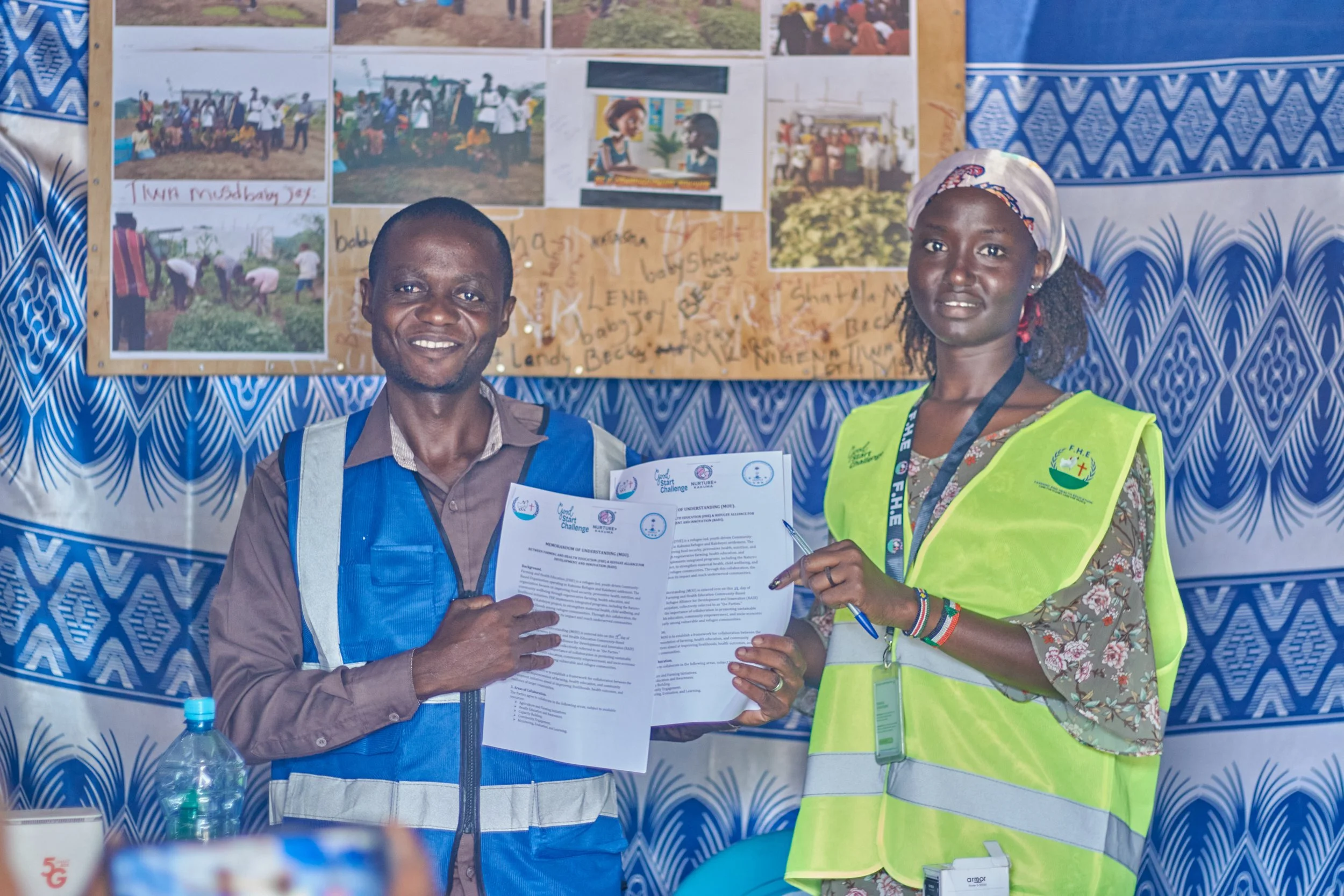 Two people, a man and a woman, are standing in front of a bulletin board with photos and writings. The man is smiling, holding papers, and wearing a blue safety vest and shirt. The woman is wearing a floral dress, a white headscarf, and a bright yell