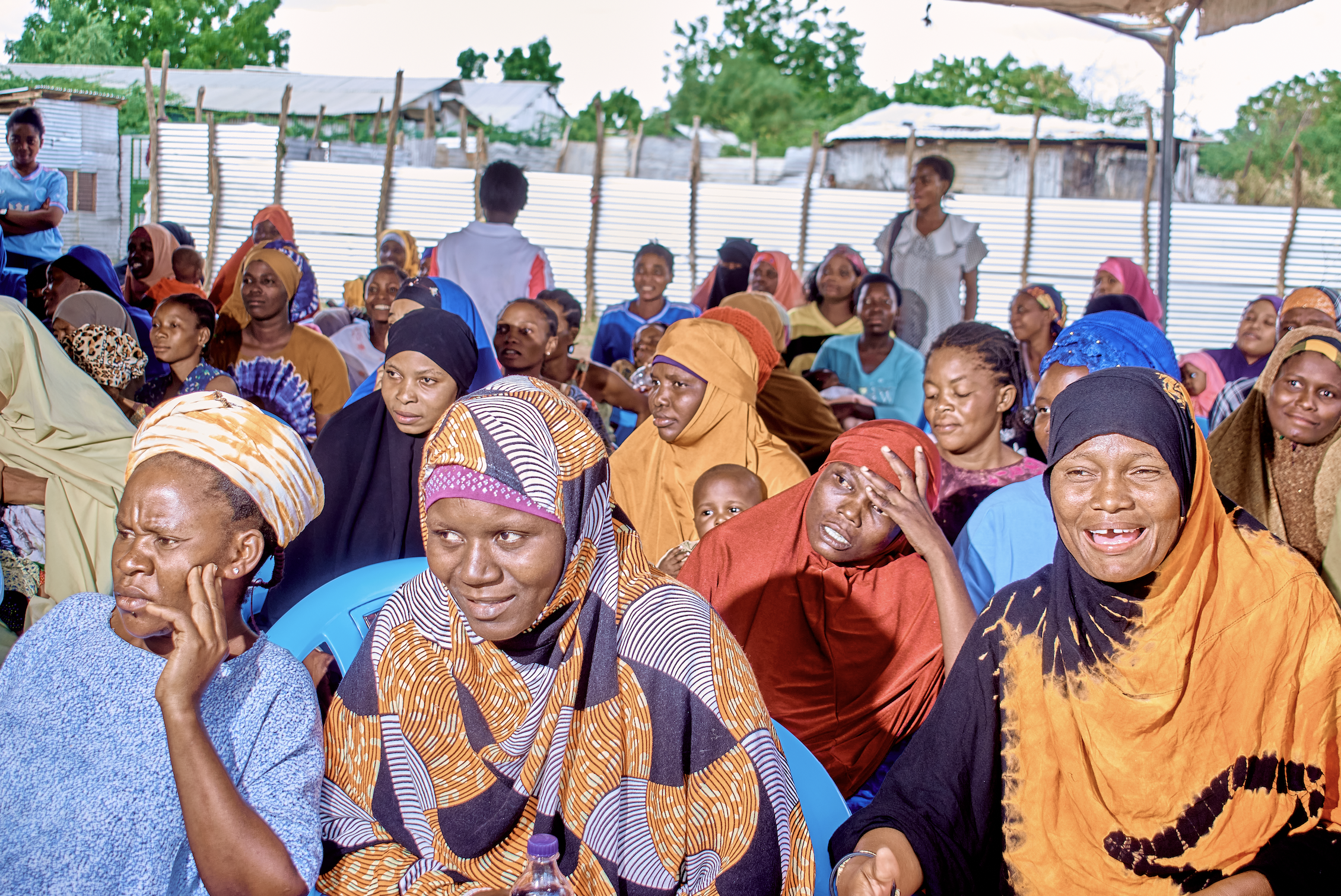 A large group of women and children gathered outdoors, some seated and some standing, under a partly covered area with makeshift fencing and trees in the background.