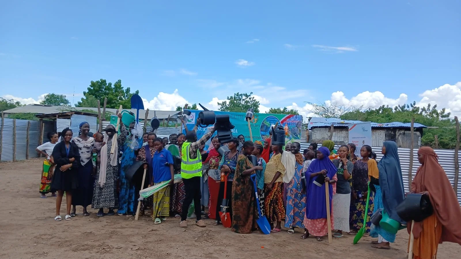 Group of people gathered outdoors, some holding shovels and gear, standing in front of a colorful banner and makeshift structures, under a cloudy sky.